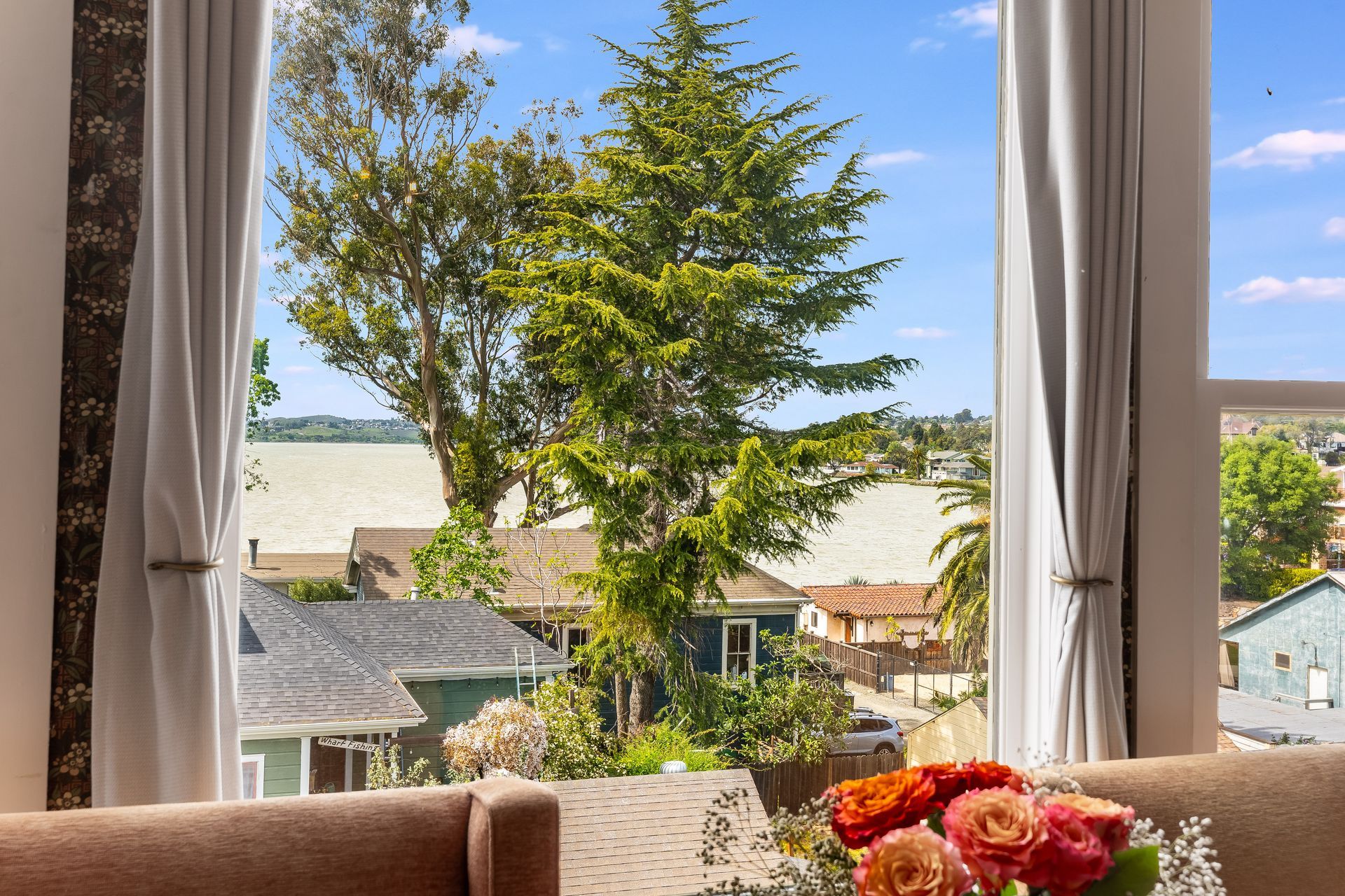 View through a window: lake, trees, houses, and a colorful flower arrangement on a table.