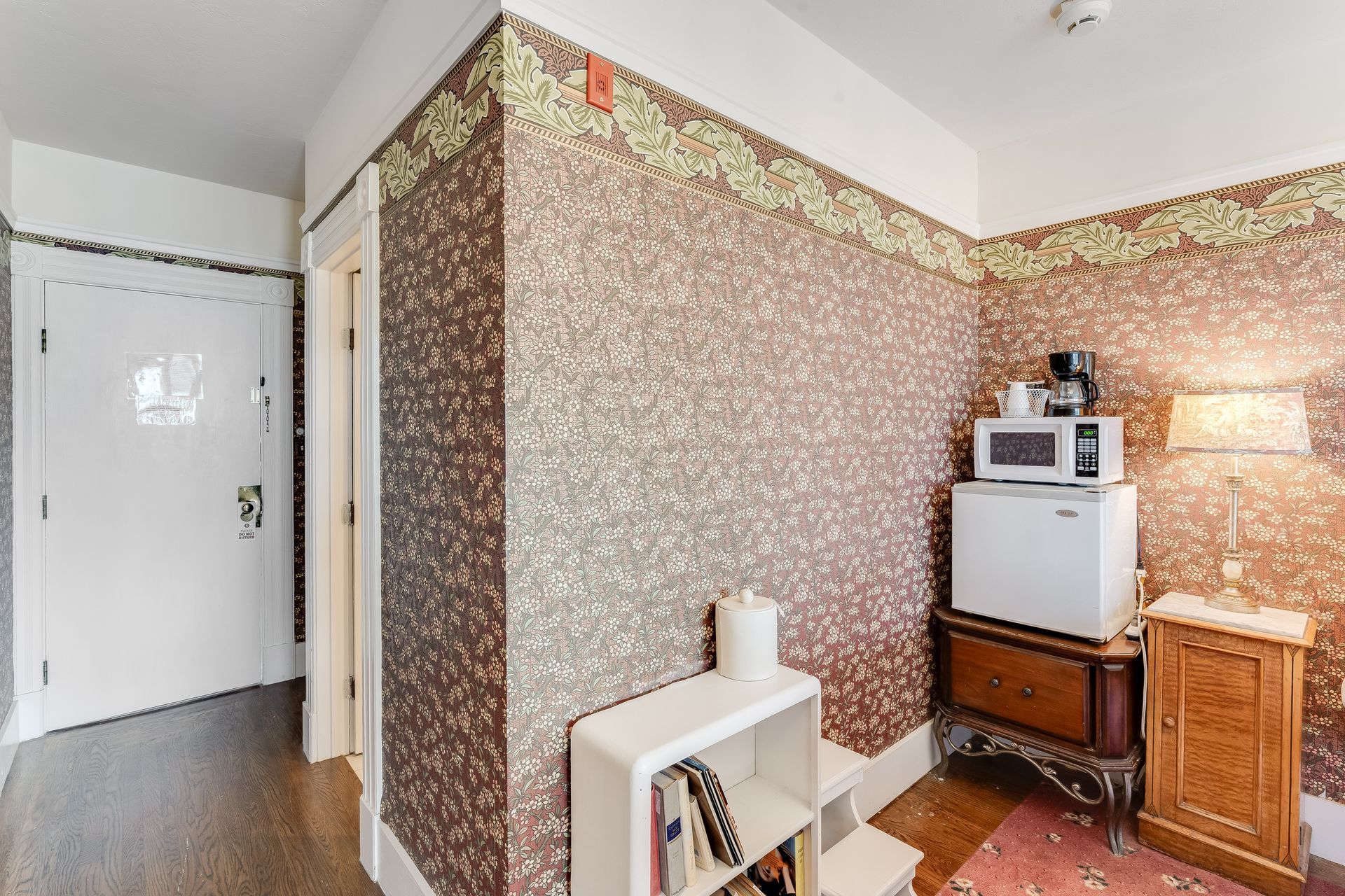 Small room with patterned wallpaper, featuring a mini-fridge, microwave, coffee maker, and nightstand.