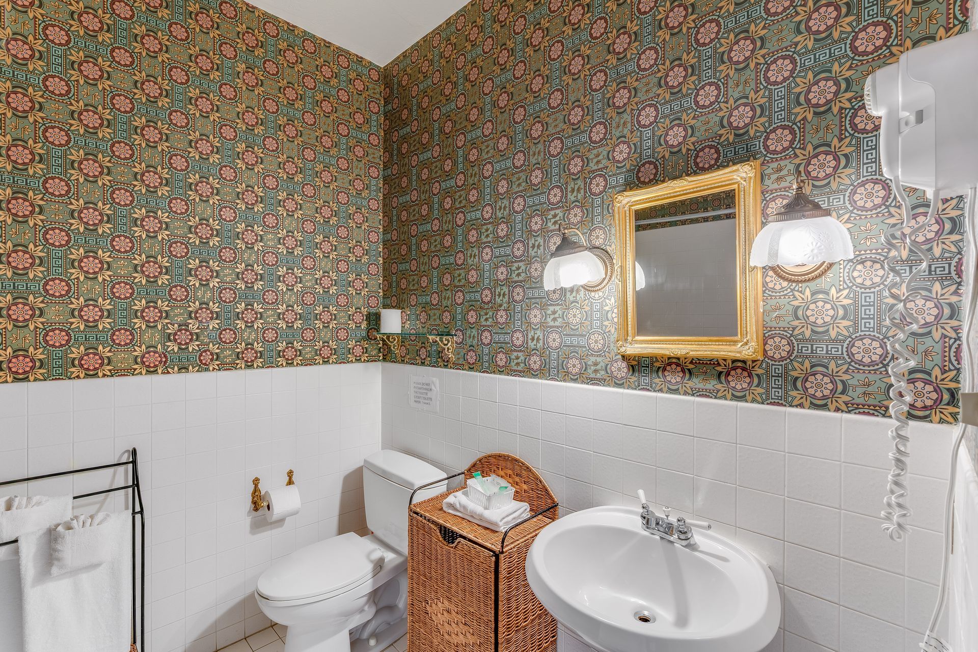 Bathroom with floral wallpaper, white paneling, gold-framed mirror, pedestal sink, and wicker hamper.
