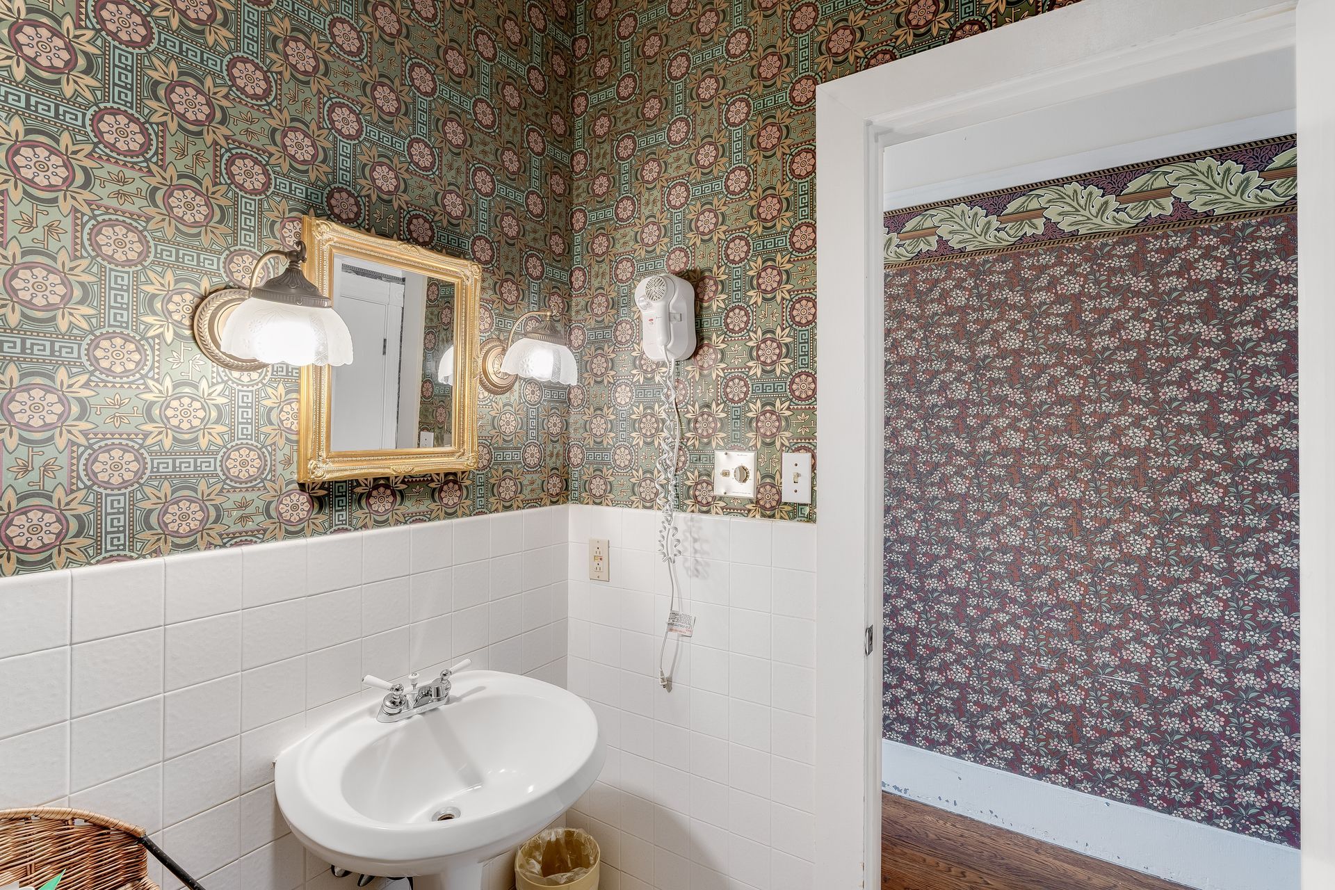 Bathroom with floral wallpaper, white sink and tiles, gold-framed mirror, and doorway to another room.
