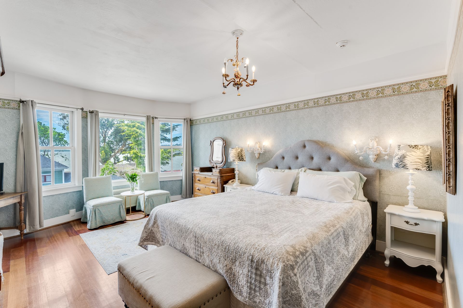 Bedroom with a large bed, curved windows, and a chandelier. Soft colors and elegant furnishings.