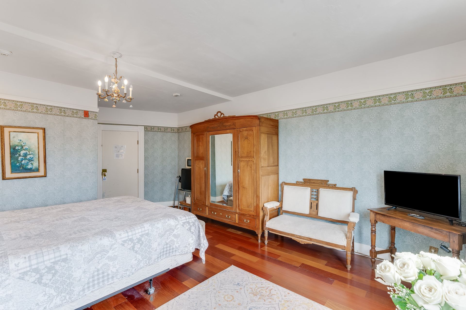 Bedroom with wooden floors, armoire, bed, small sofa, television, and floral wallpaper.