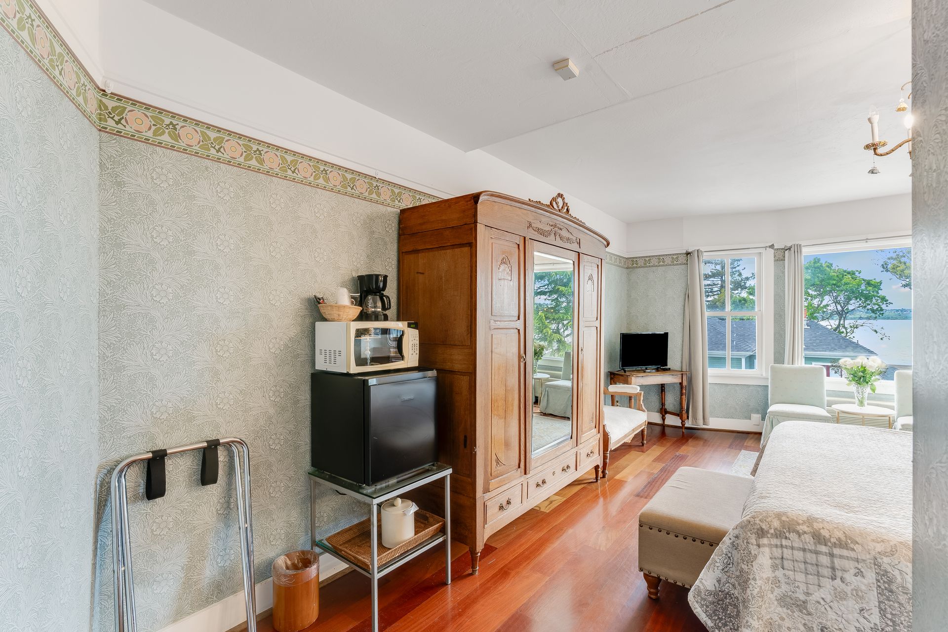 Hotel room with antique wardrobe, microwave, mini-fridge, luggage rack, small table, TV, and a view of the water.