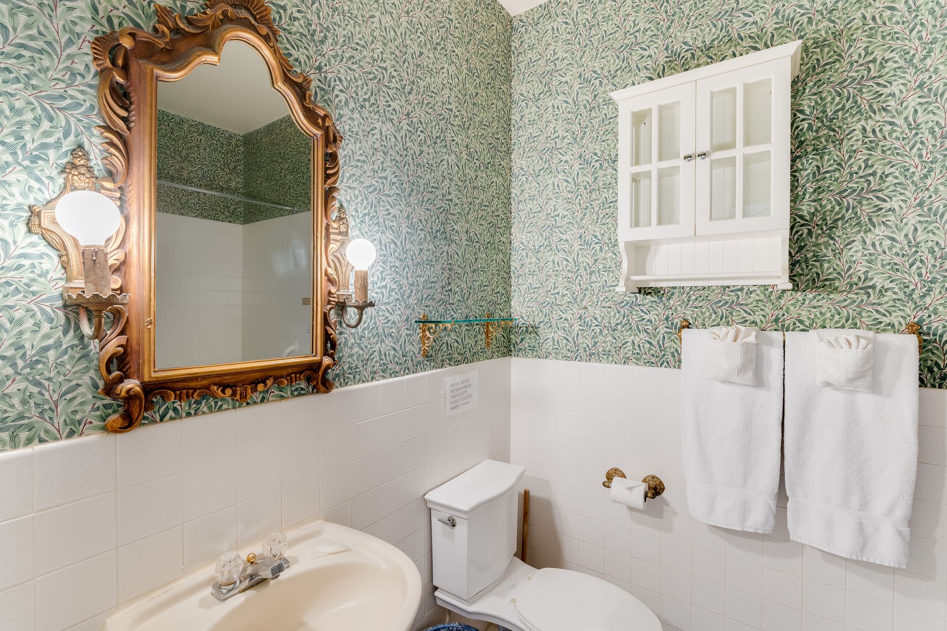 Bathroom with ornate gold mirror, patterned wallpaper, white fixtures, and towels.