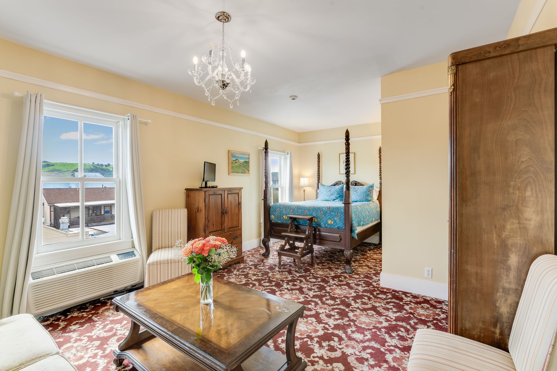 Hotel room with a four-poster bed, seating area, window overlooking a harbor, floral carpet, and chandelier.