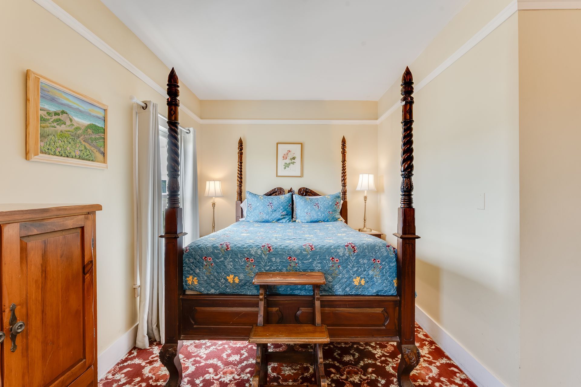 Bedroom with dark wood four-poster bed, blue floral bedding, and wooden step stool on red patterned rug.