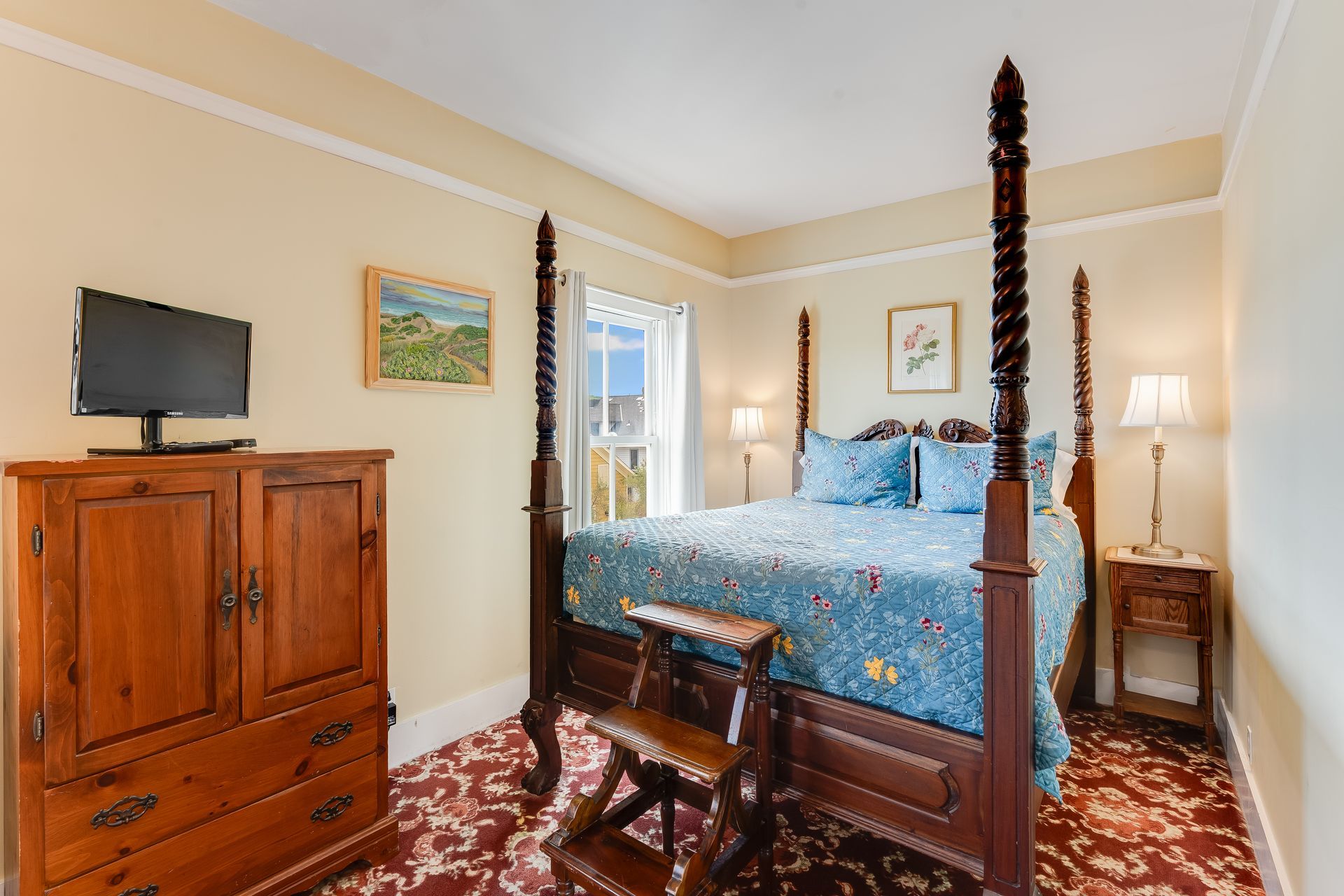 Bedroom with four-poster bed, TV, and floral bedspread; wood furniture and rug, light yellow walls.