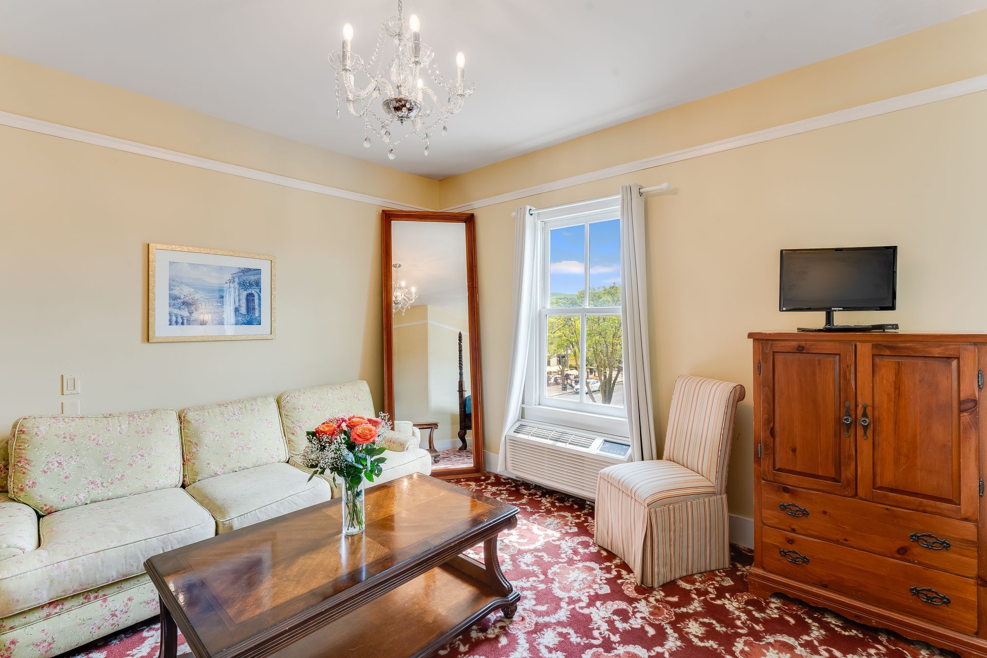 Living room with cream sofa, wooden furniture, floral rug, and a window with a view.