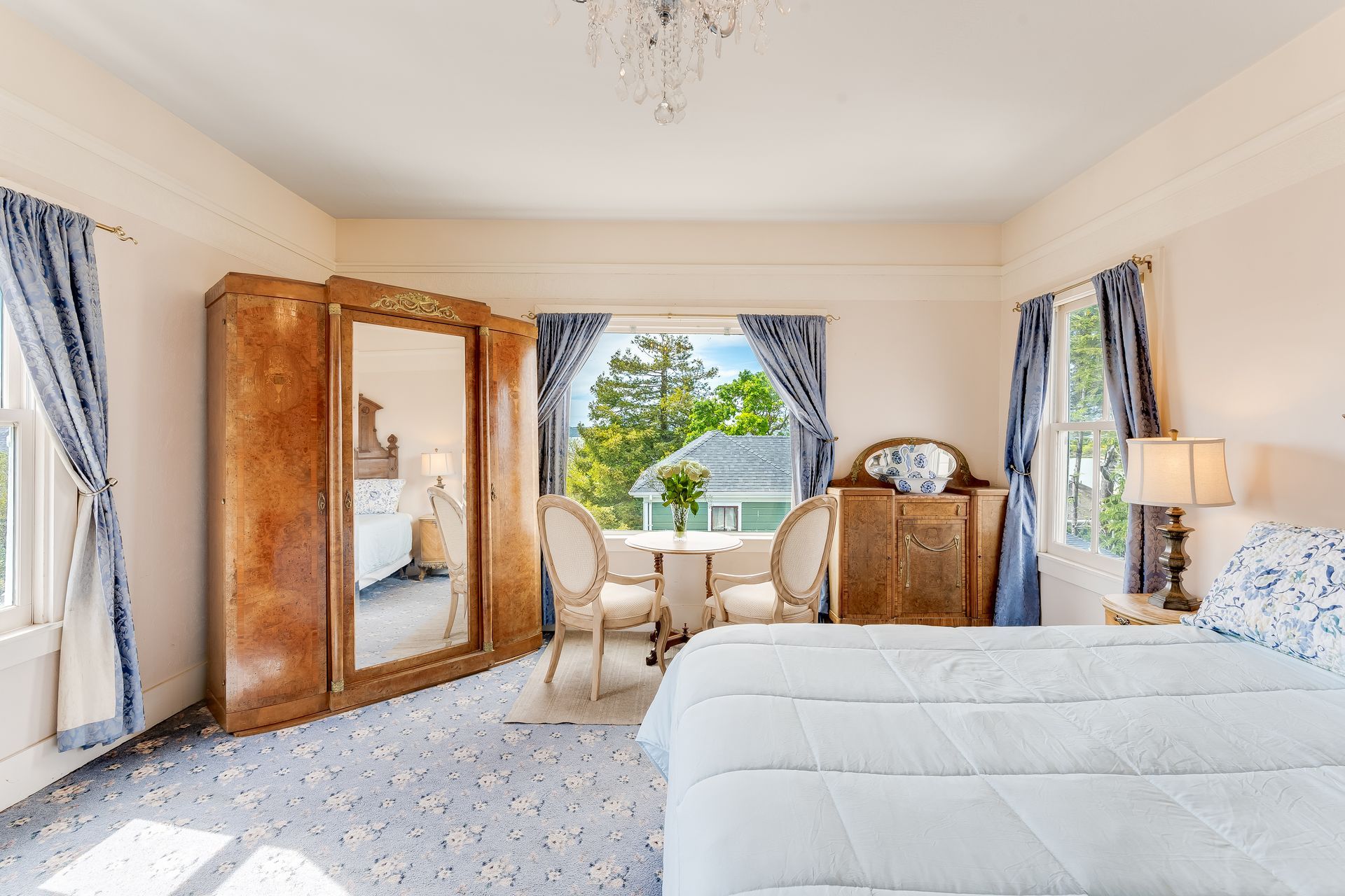 Bedroom with bed, wardrobe, small table, and windows with blue curtains.