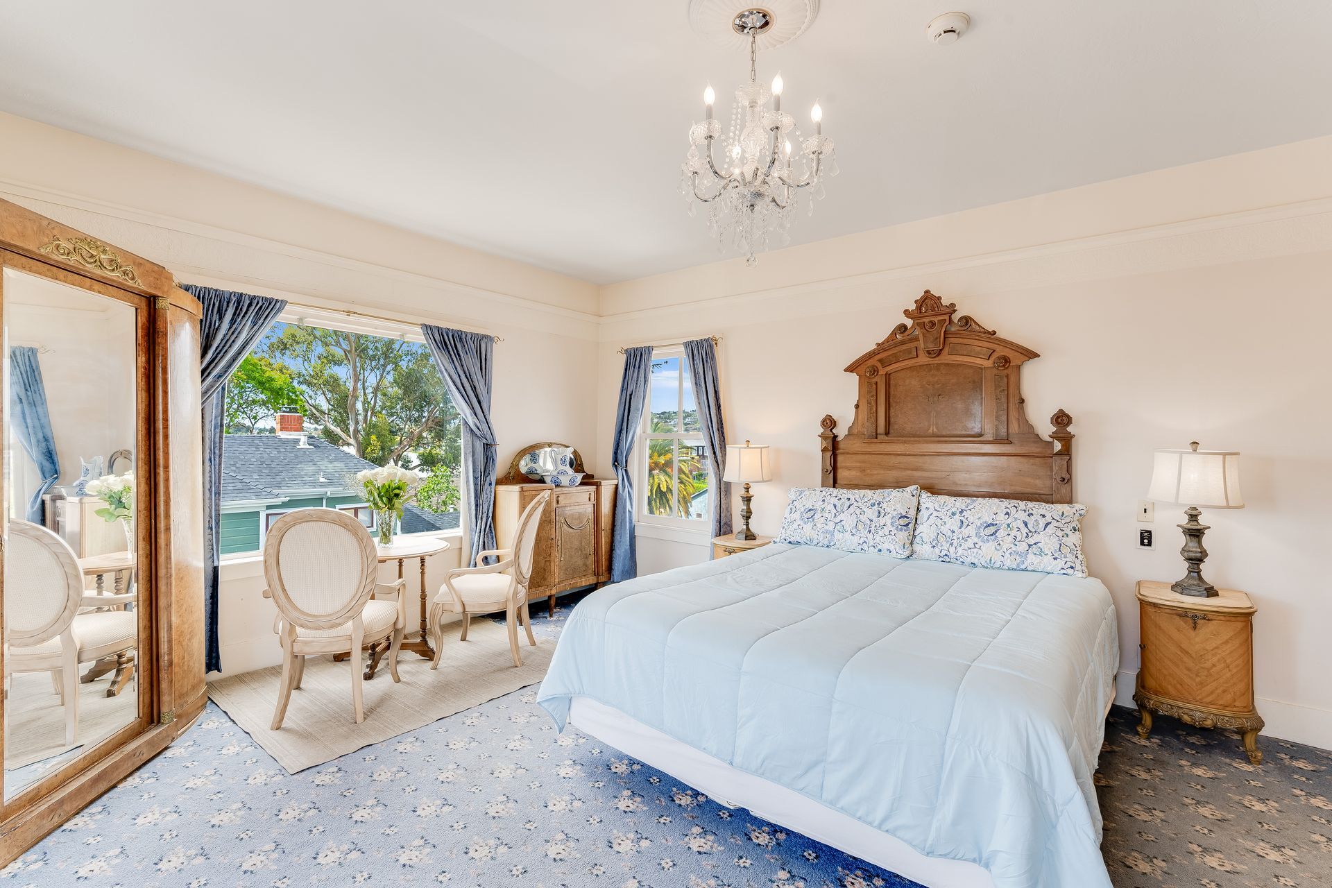Elegant bedroom with light blue bedding, ornate wooden headboard, chandelier, and window with a view.