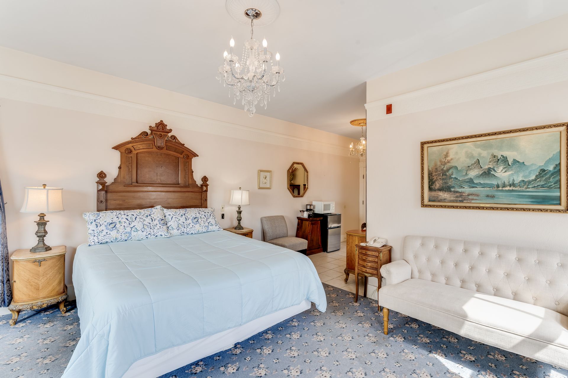 Bedroom with ornate wooden headboard, blue accents, chandelier, sofa, and artwork.