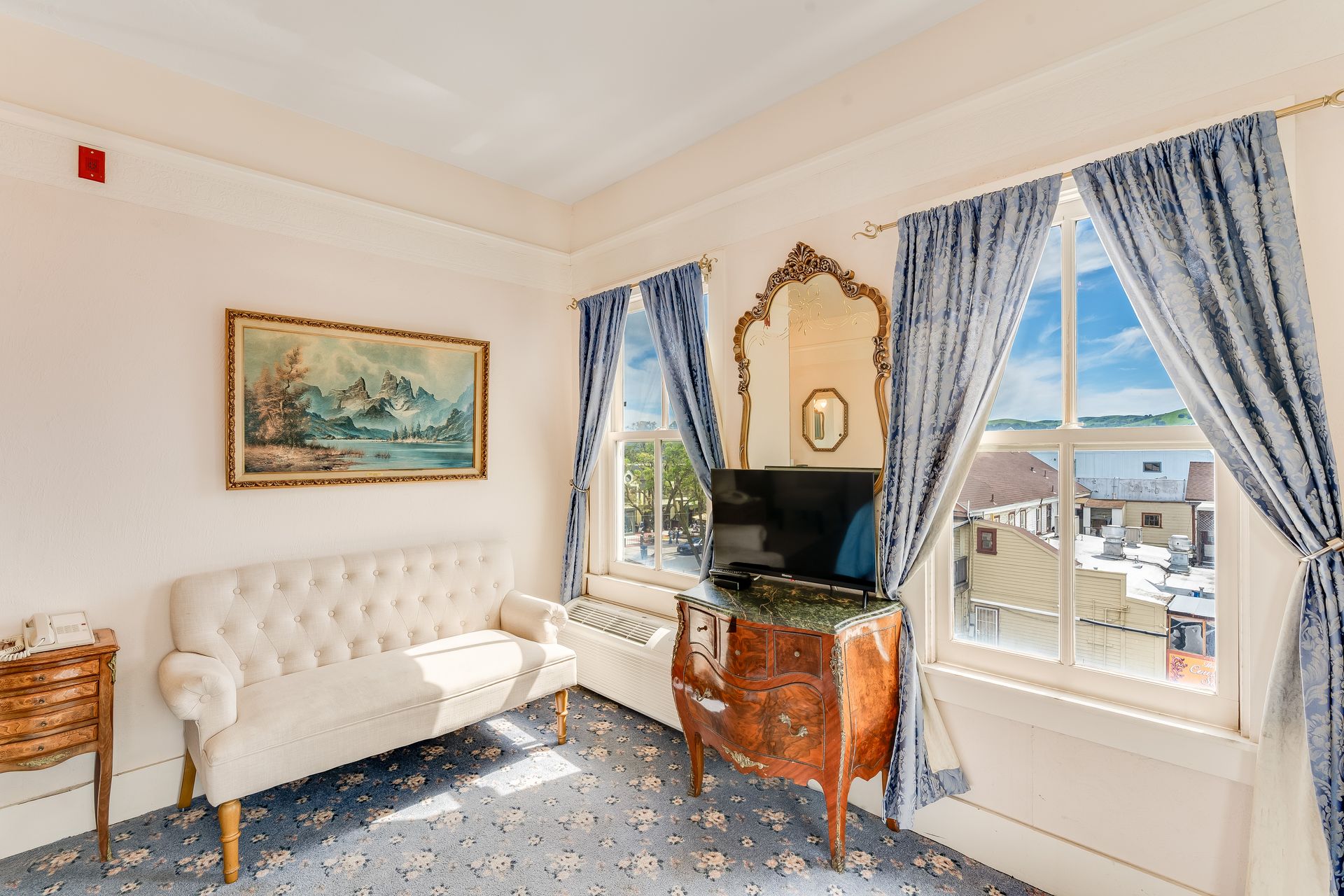 A light-filled hotel room with a white couch, two windows, and a TV on a decorative stand.