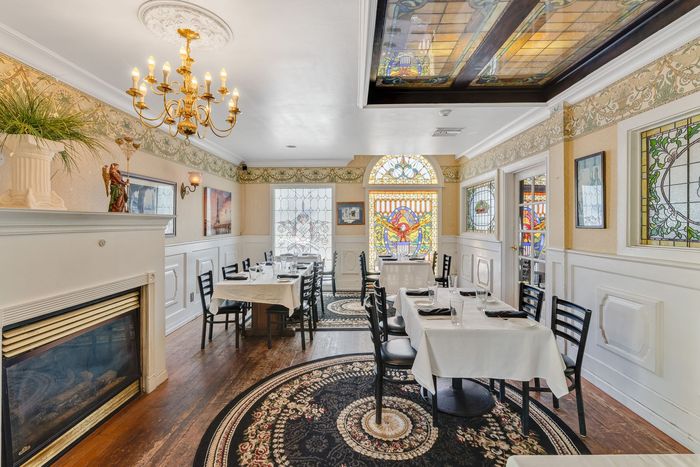 Restaurant interior: tables with white linens, black chairs, fireplace, stained glass windows, chandelier, patterned carpet.