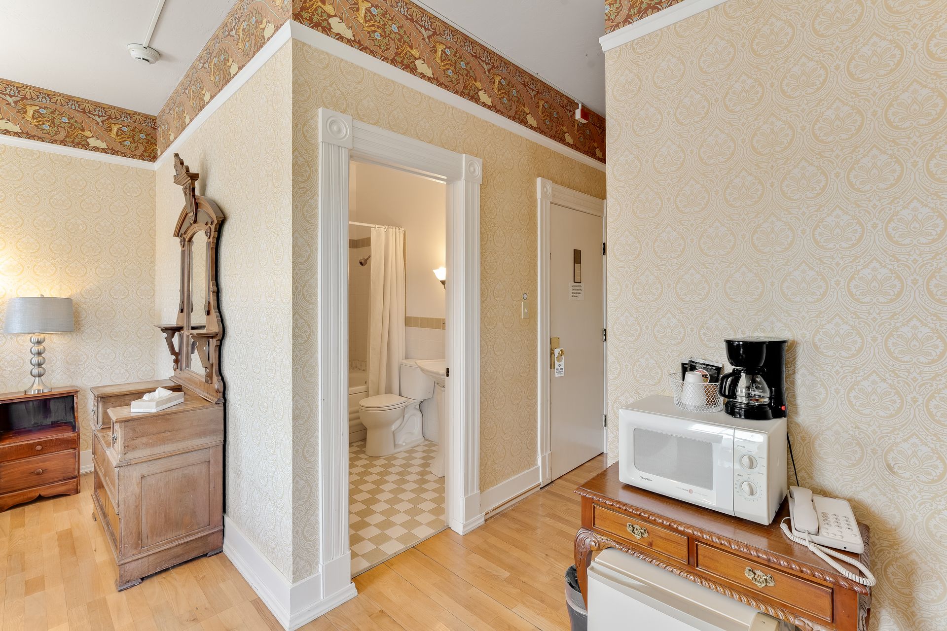 Hotel room hallway with a bathroom, microwave, dresser, and mirror. Light walls, wood floors.