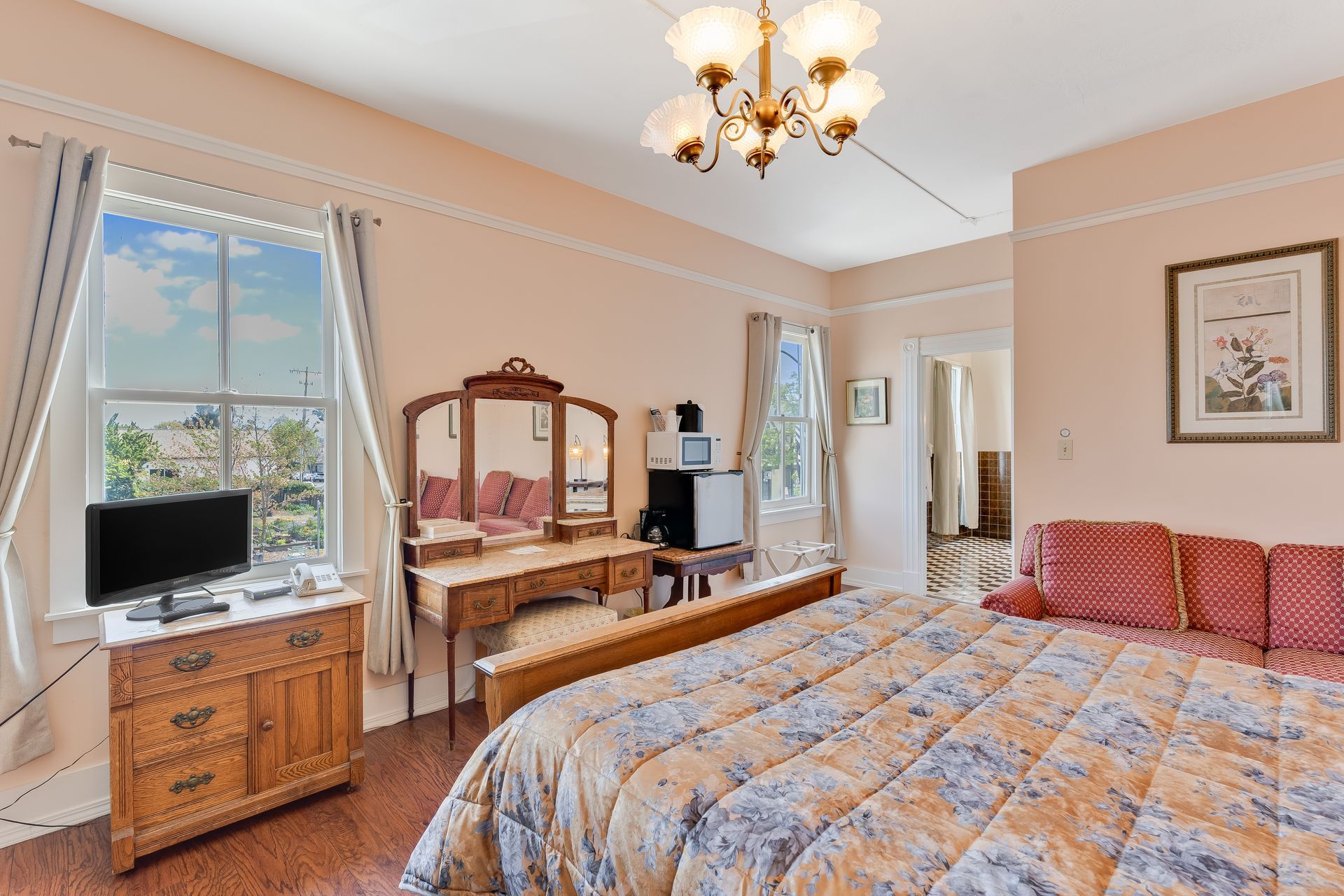 Bedroom with antique furniture, including a vanity, dresser, and bed. Pink walls, large windows, and chandelier.