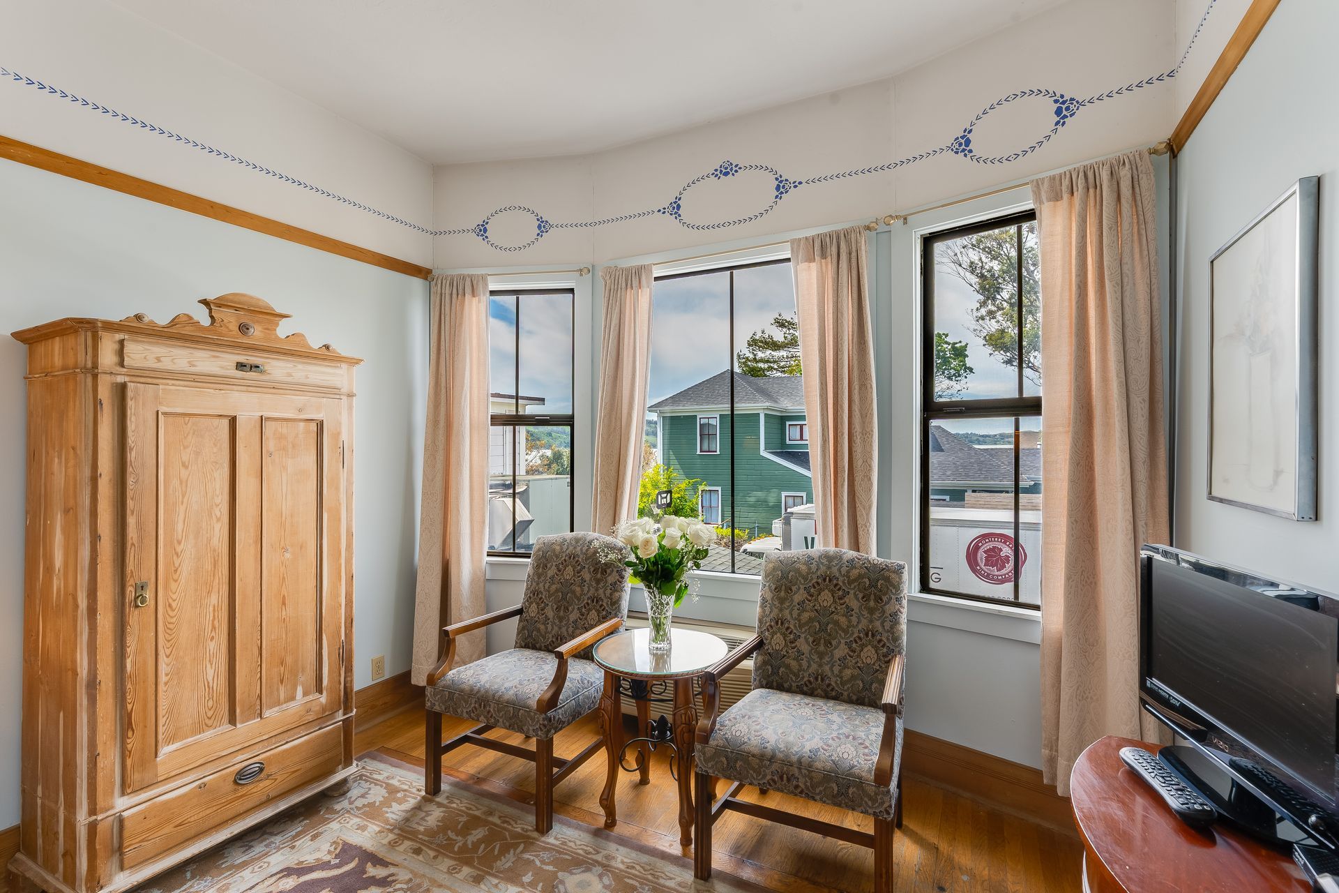 Bedroom with bay window, antique armoire, two chairs, small table, floral painting.