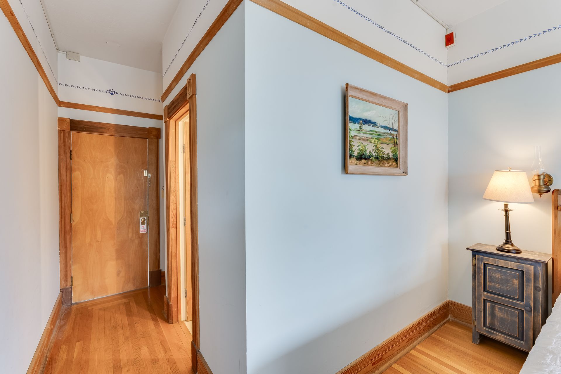 Hallway with wood floors, pale blue walls, wooden trim, and a nightstand with a lamp.