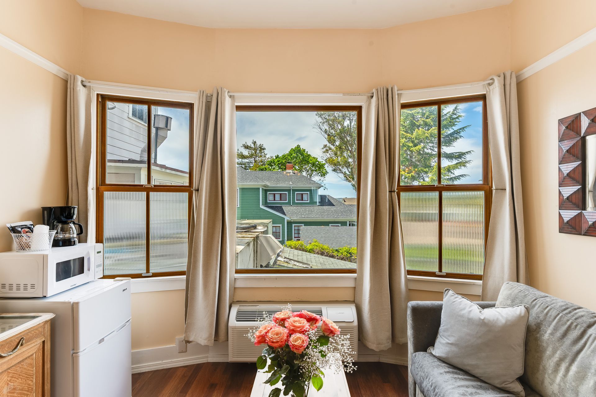 Cozy room with bay window, beige curtains, sofa, and mini-fridge, overlooking a residential neighborhood.