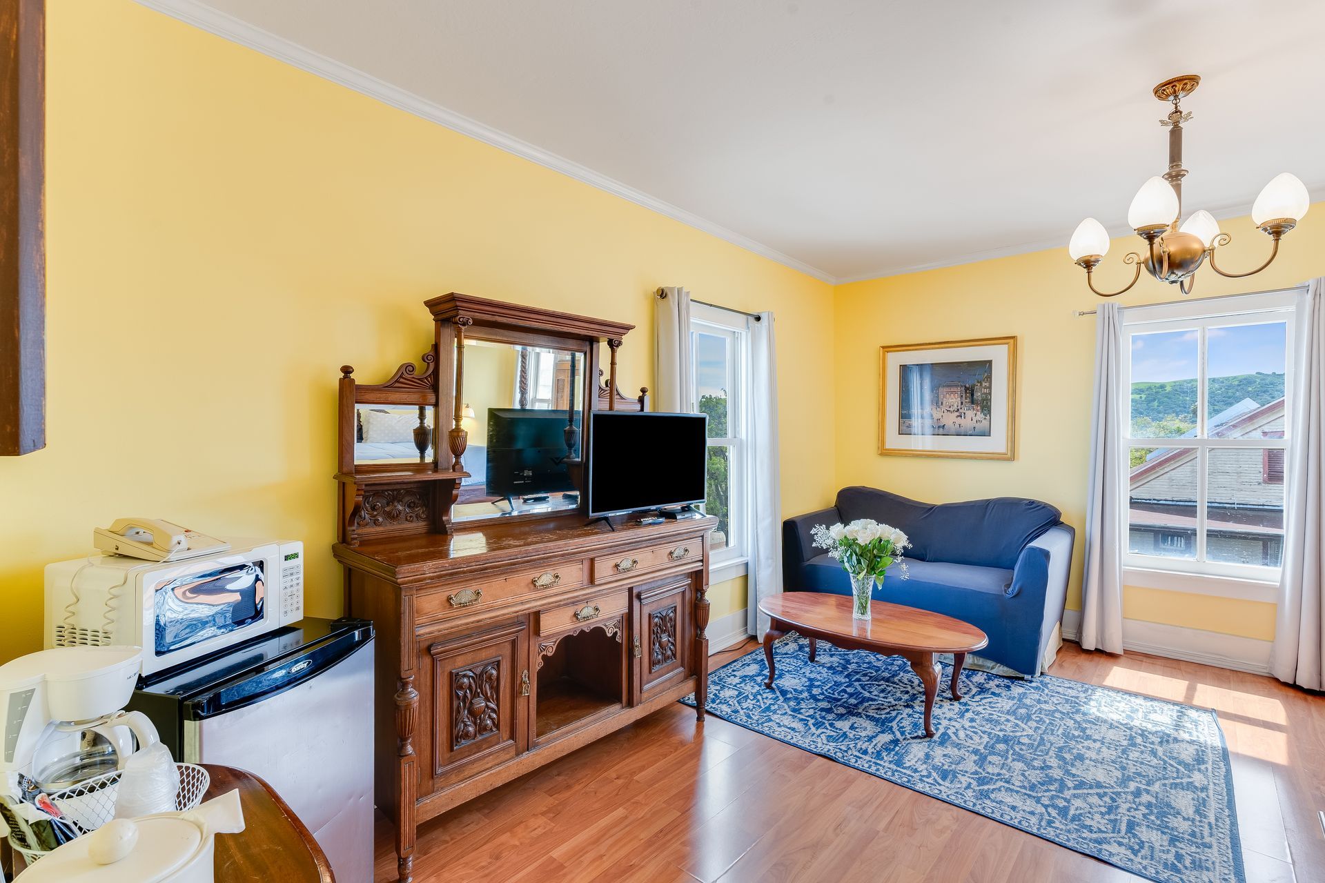 Yellow-walled room with dark wood furniture, blue couch, and a small rug. Two windows, a painting, and a chandelier.