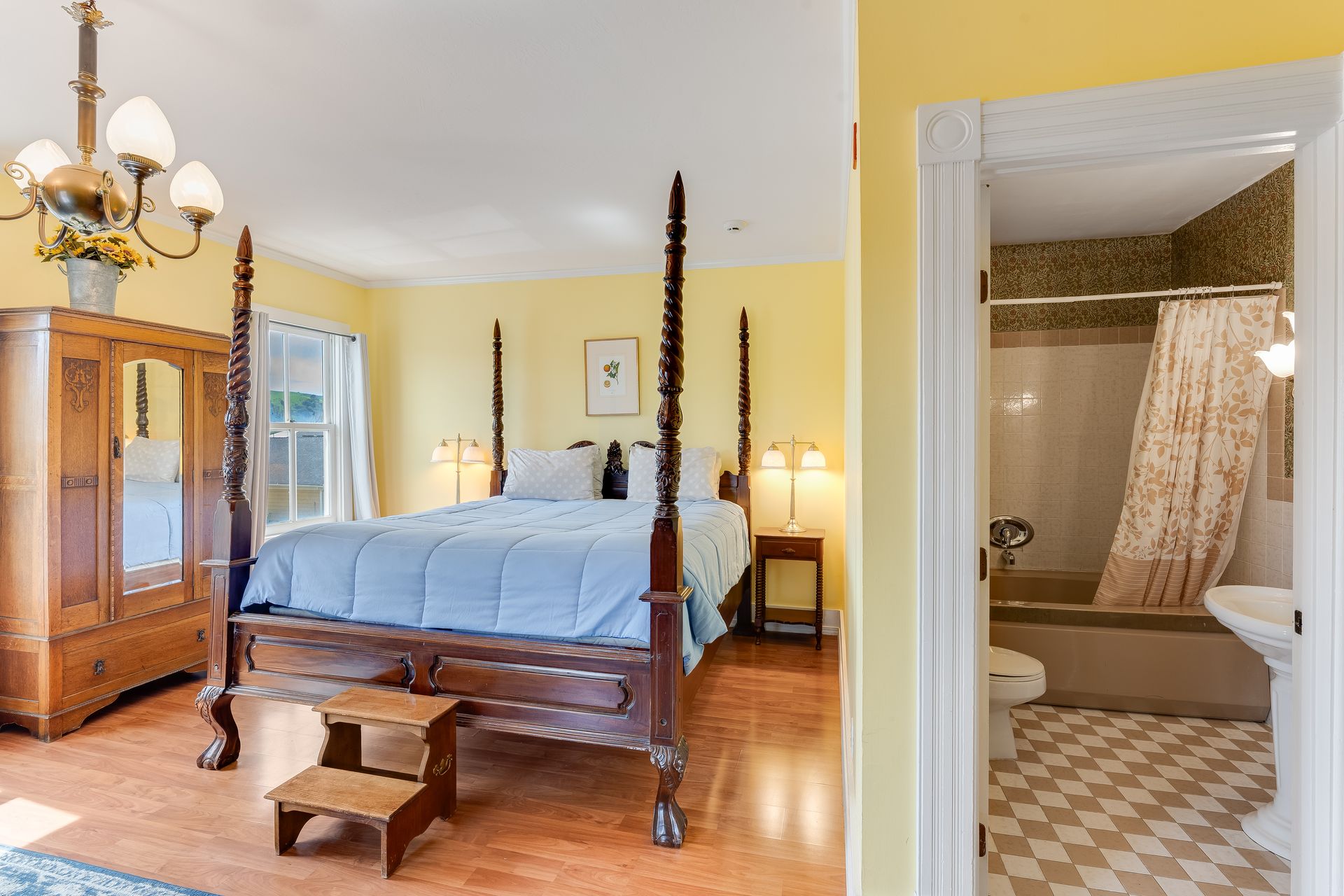 Bedroom with four-poster bed, antique wardrobe, and bathroom visible through doorway. Yellow walls, hardwood floors.