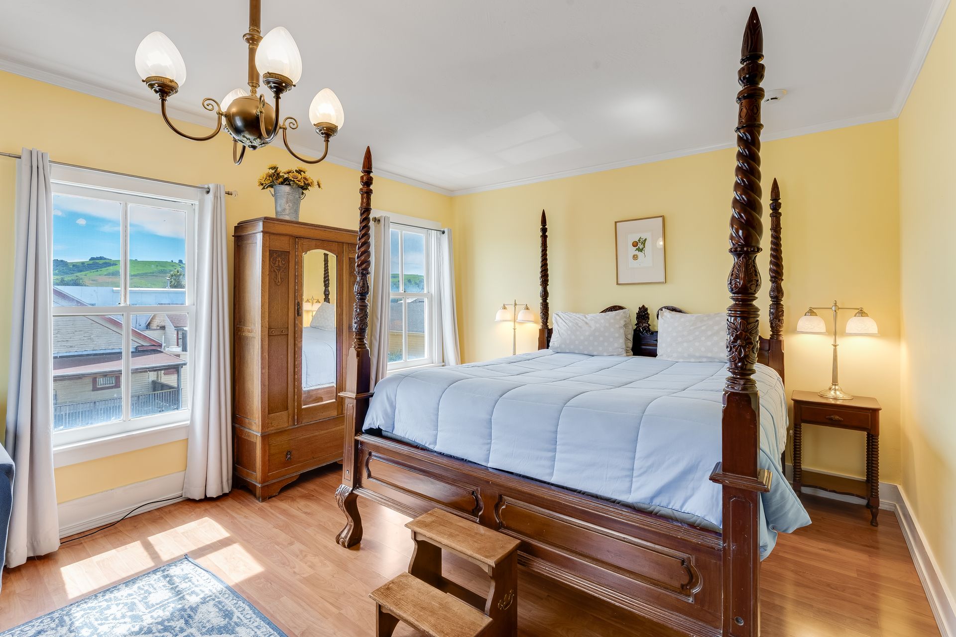Yellow bedroom with four-poster bed, wooden wardrobe, chandelier, and two windows overlooking a rural view.