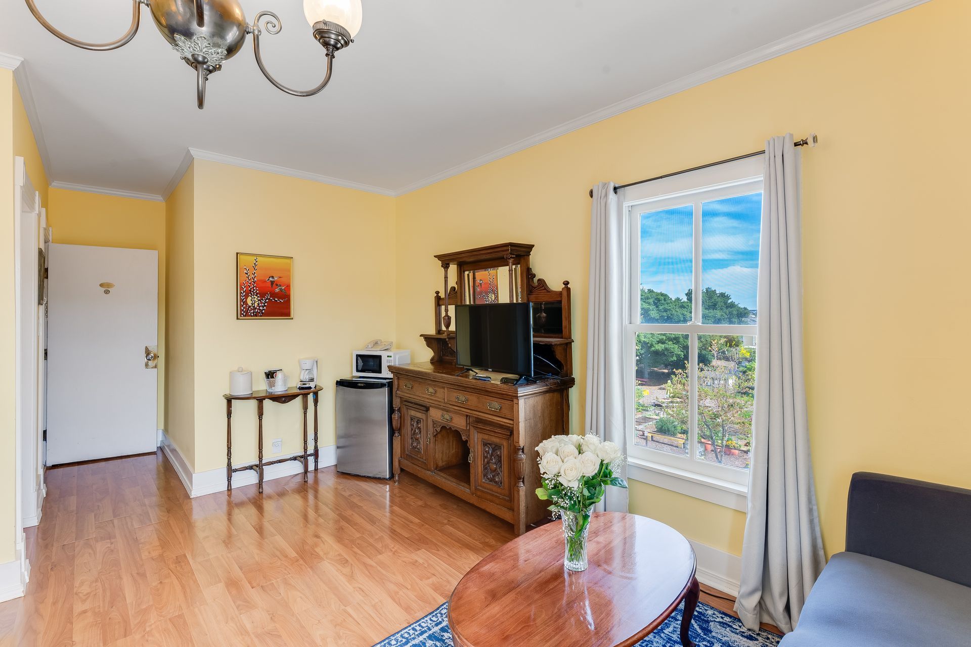 Yellow room with wooden furniture, a window with a view, and a bouquet of white flowers.