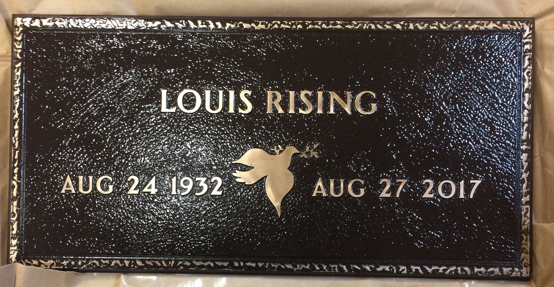 A black, rectangular grave marker with a gold-colored border, inscribed: 