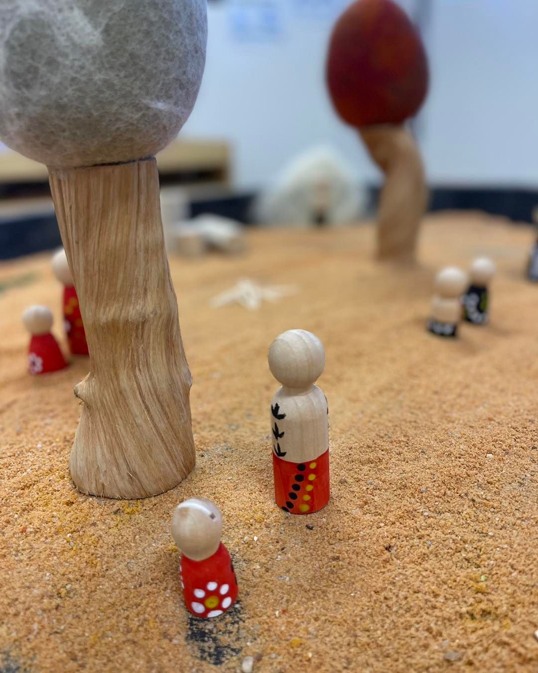 Wooden peg dolls in a sandy play area with felt trees; some dolls wearing red and black clothing.