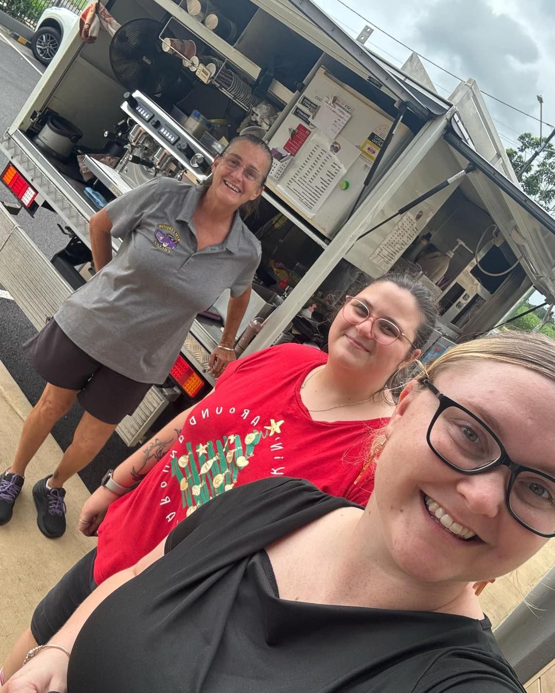 Three women smiling near a food truck. Two are in front, one is behind, working inside.