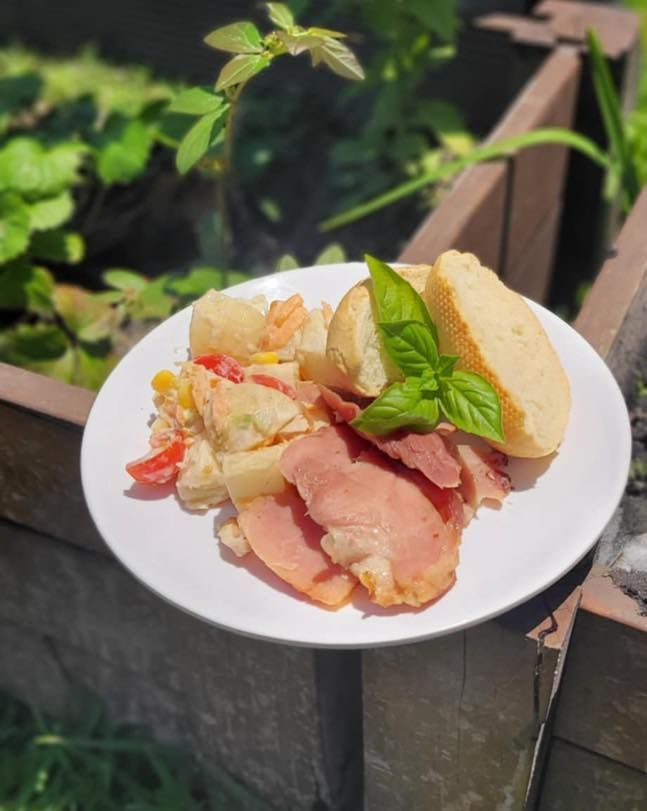 Plate with salad, ham, and bread, garnished with basil. Outdoors, garden in background.
