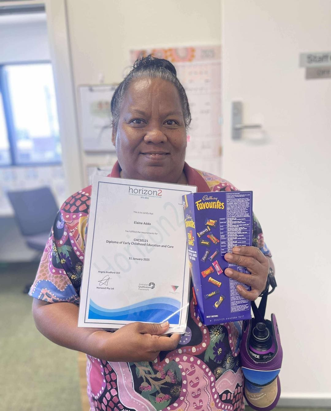 Woman holding certificate and box of chocolates, smiling indoors.