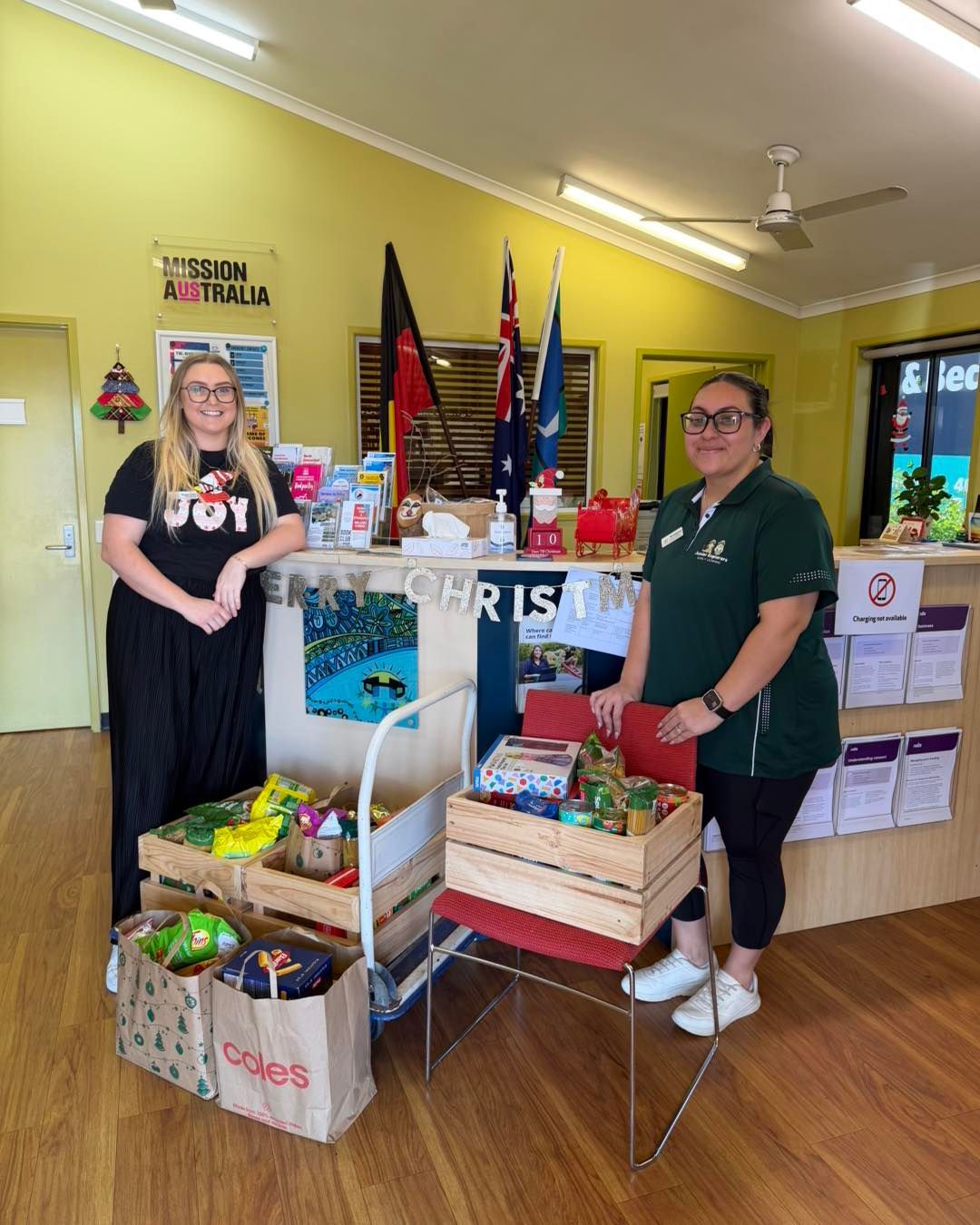 Two women standing behind a counter with food donations. Yellow wall, flags, and Coles bag visible.