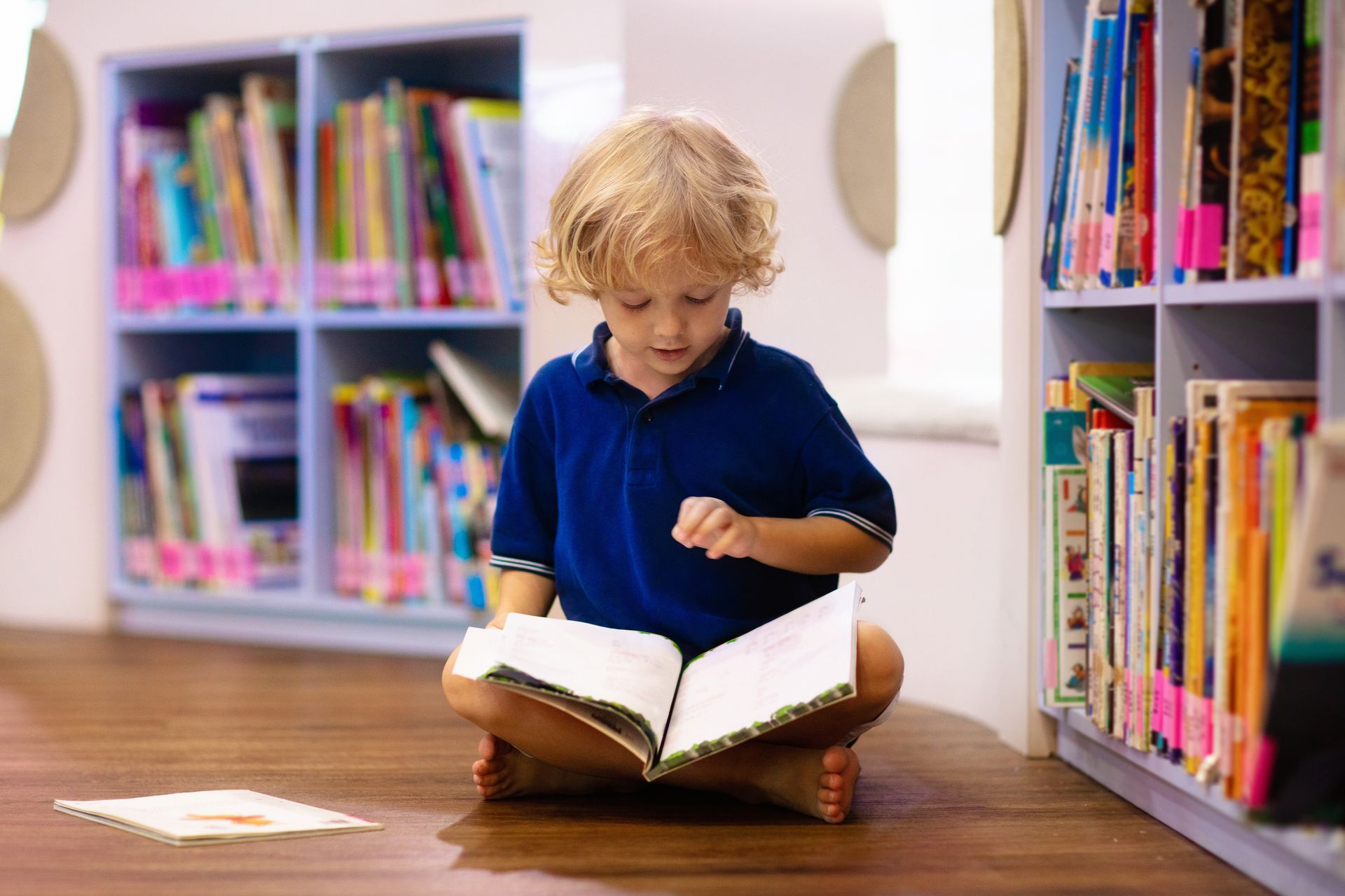 Young child with blond hair sits cross-legged, reading a book on a library floor, surrounded by bookshelves.