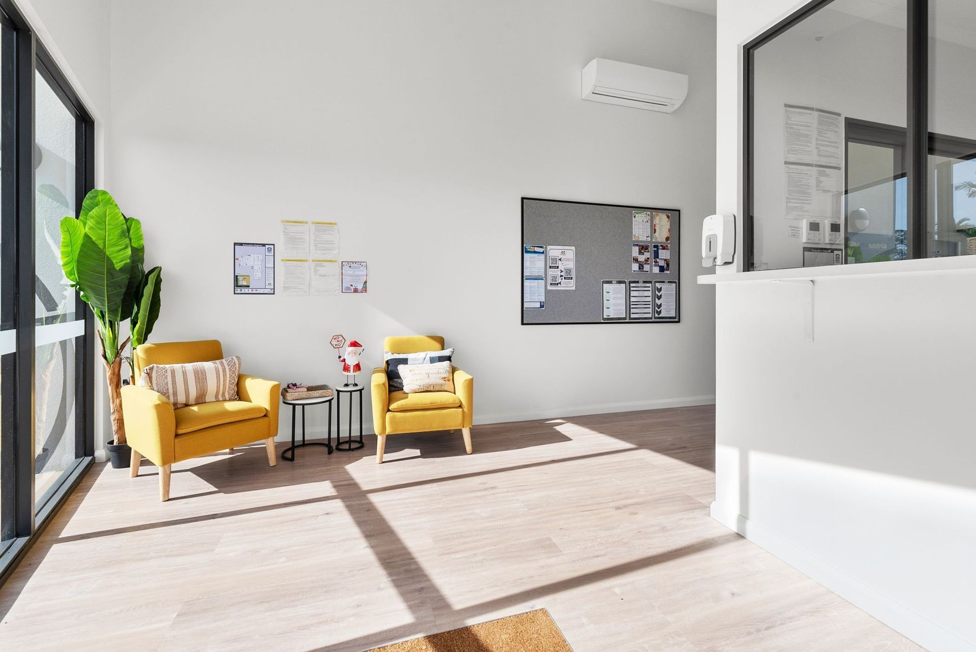 Bright waiting room with yellow armchairs, plant, corkboard, and reception window.
