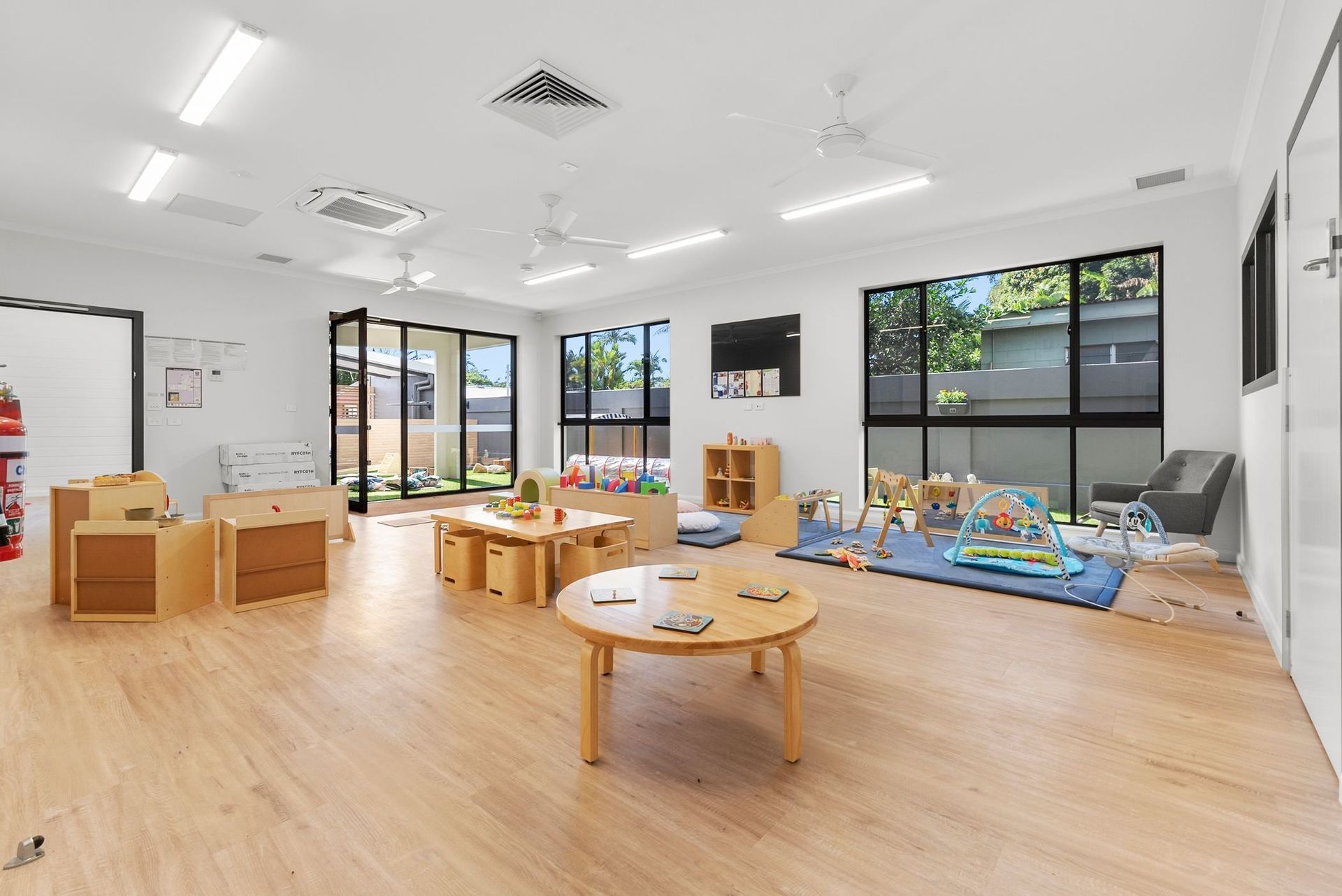 Brightly lit playroom with wooden tables, toys, and large windows.
