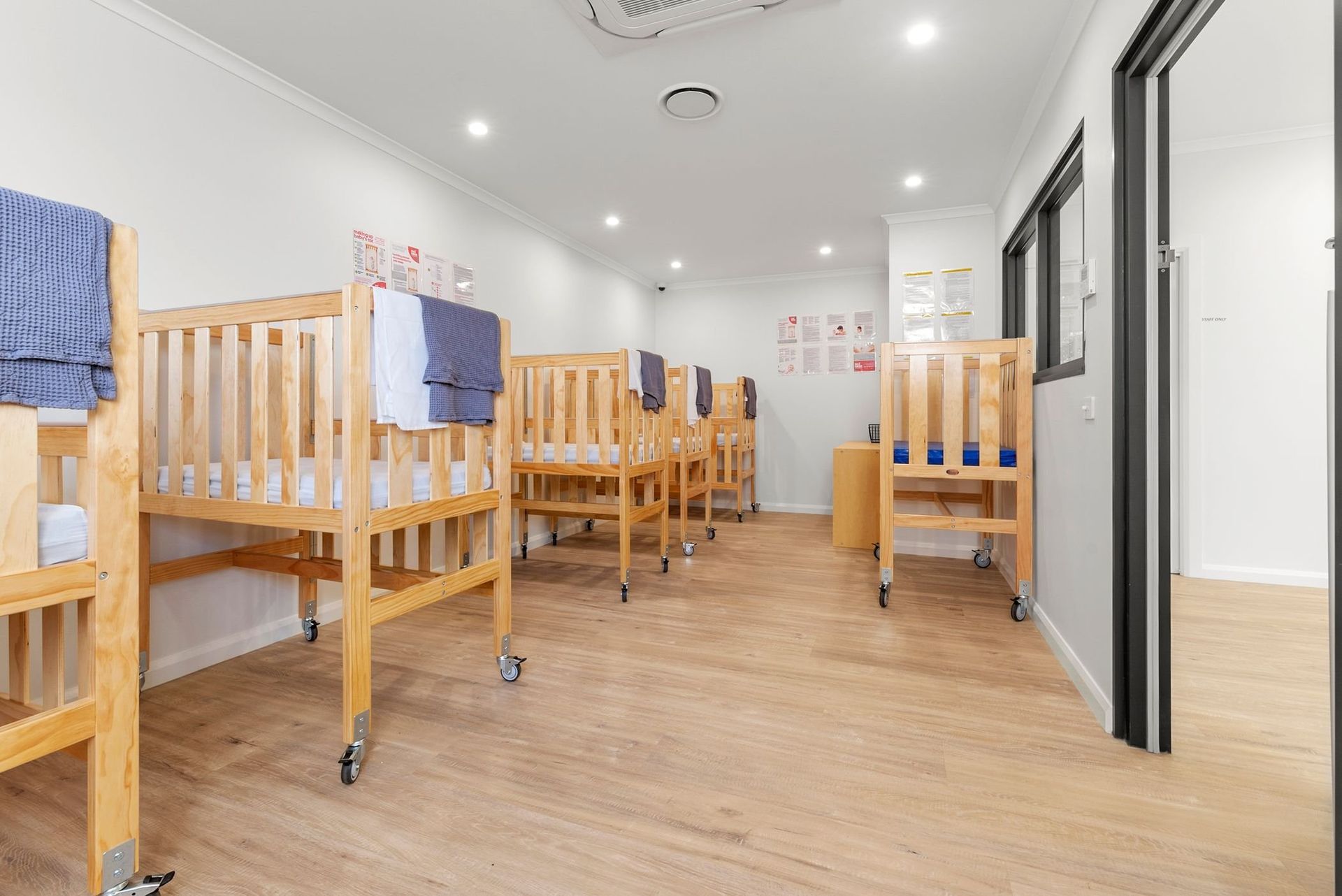 Nursery room with wooden cribs on wheels, white walls, and a light wooden floor.