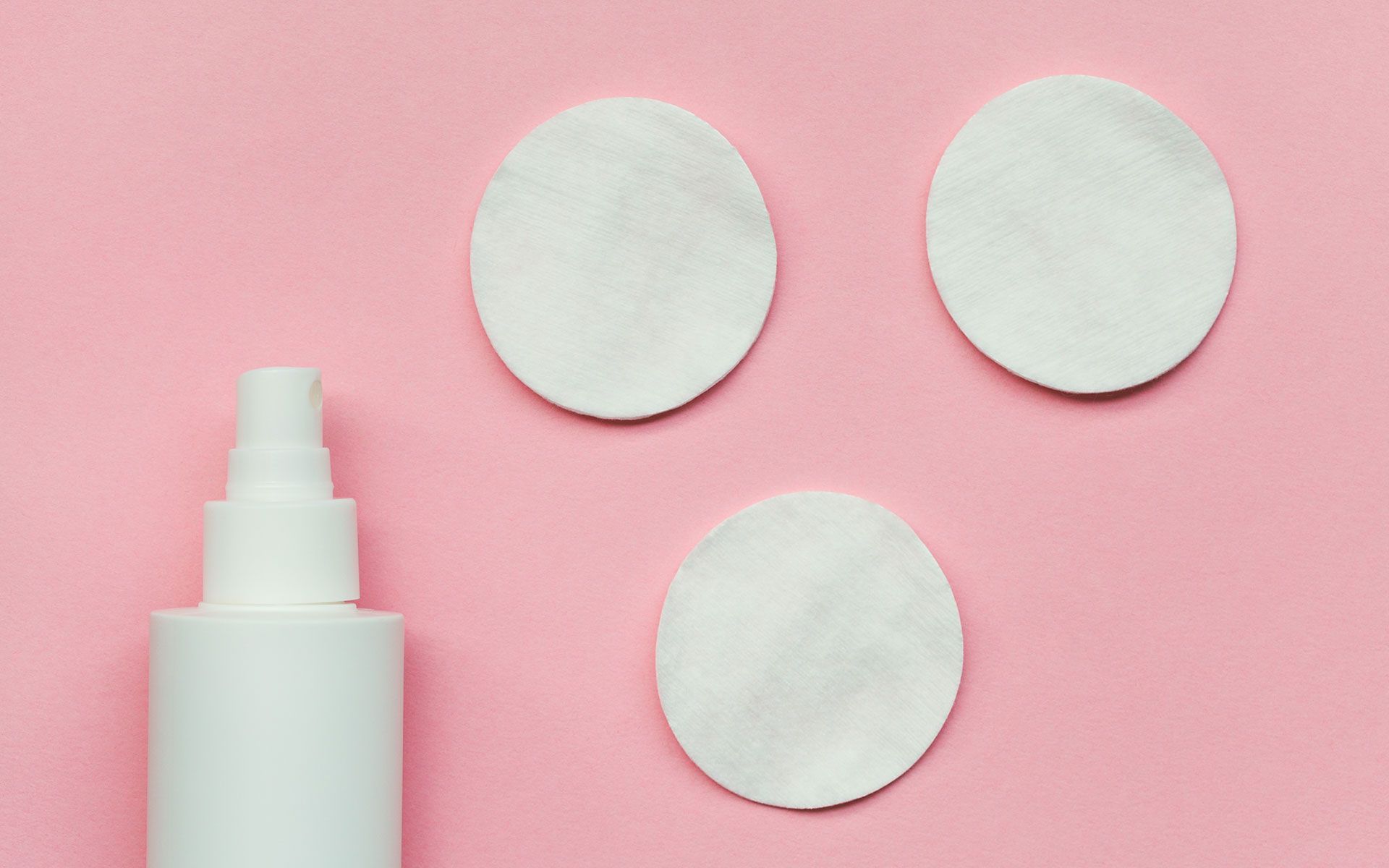 White spray bottle and cotton pads on a pink background.