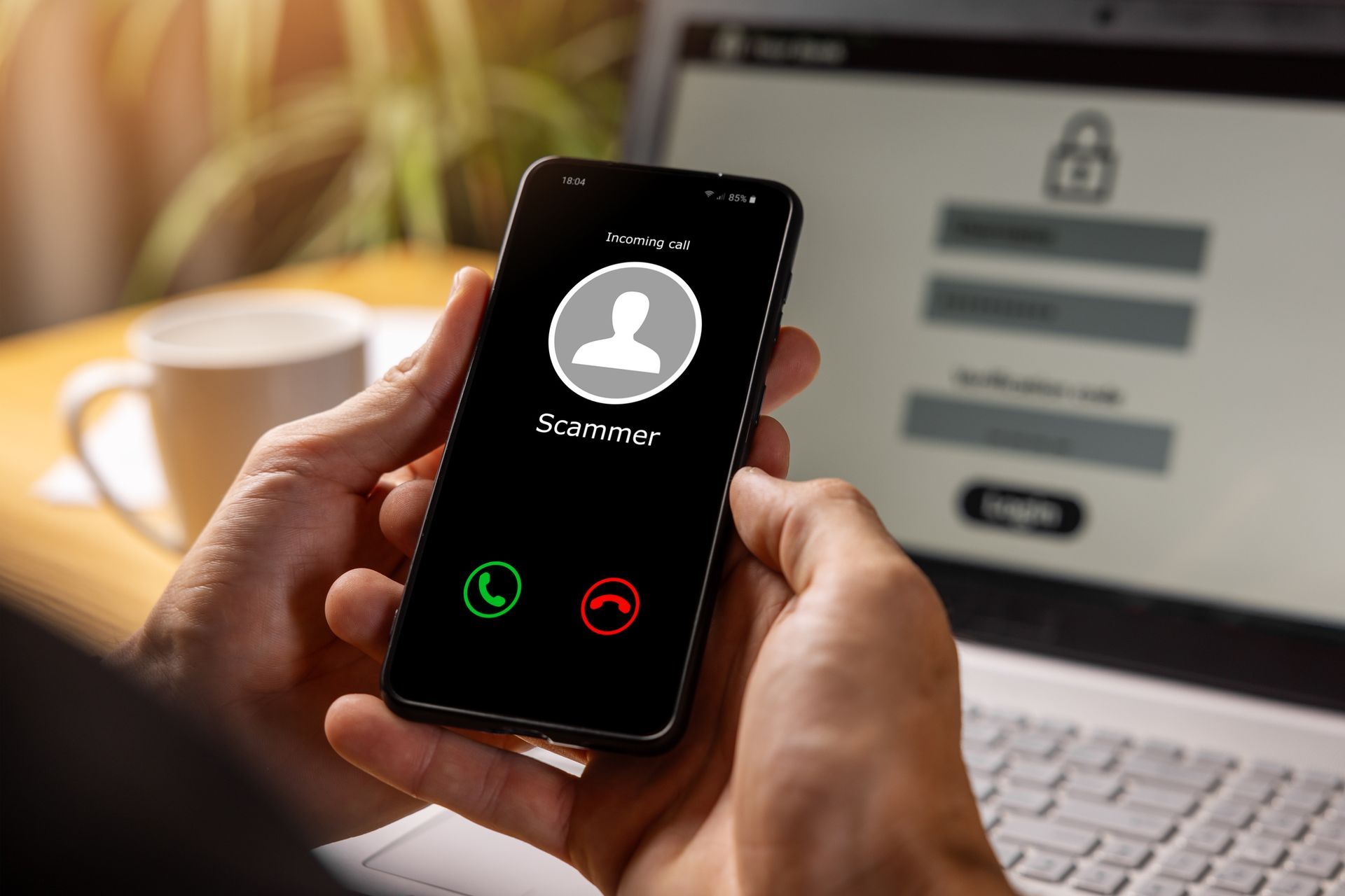 Hand holding smartphone showing an incoming call screen beside a laptop with a login page