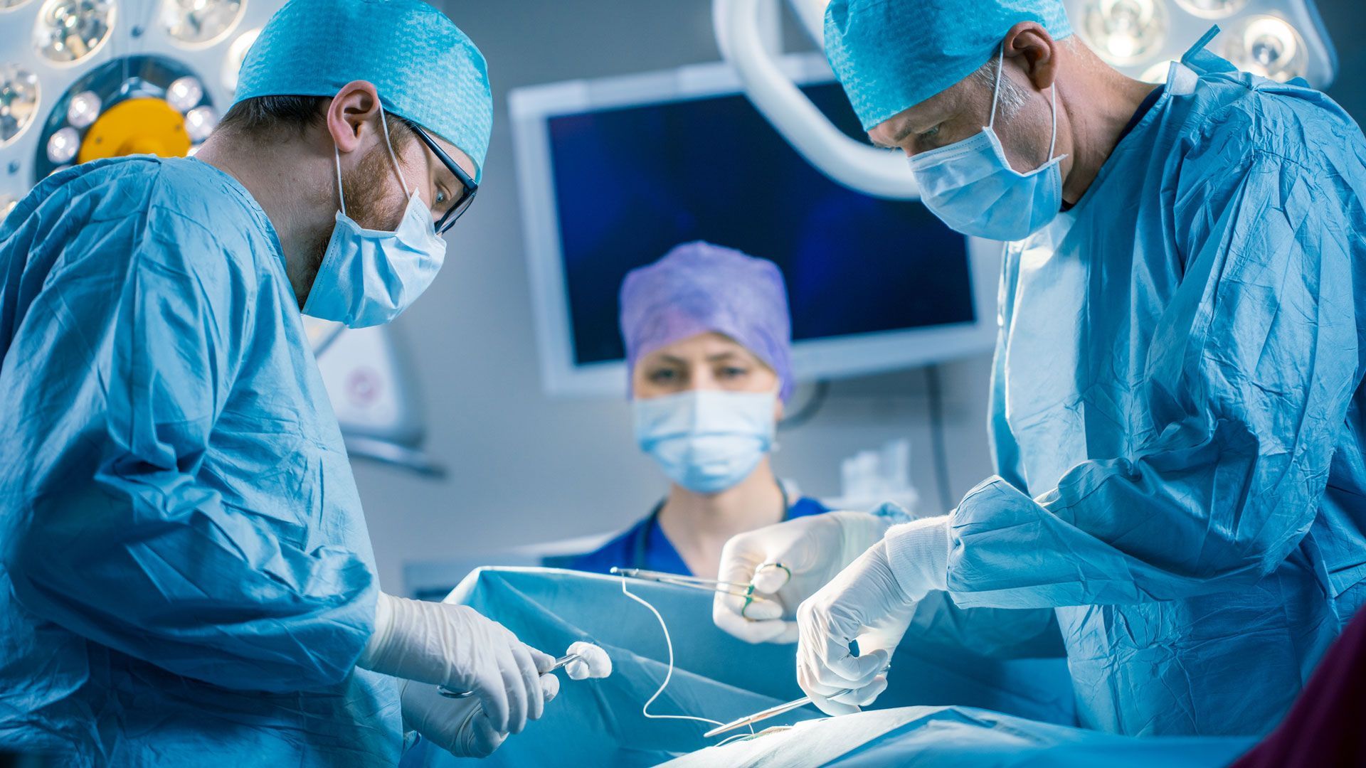 Surgeons in blue scrubs and masks perform an operation under bright operating-room lights.