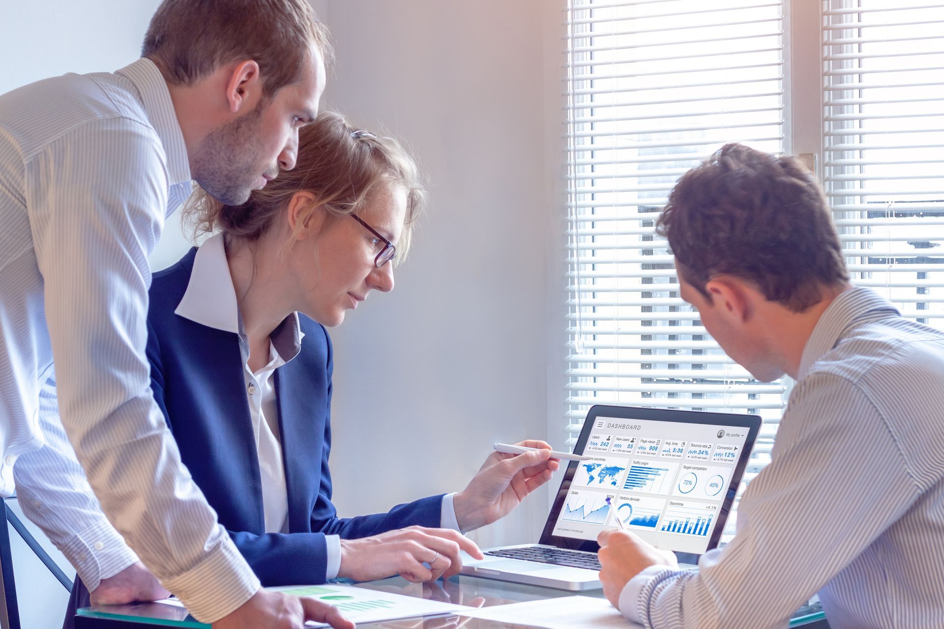 Three coworkers discuss data on a laptop in a bright office meeting room.