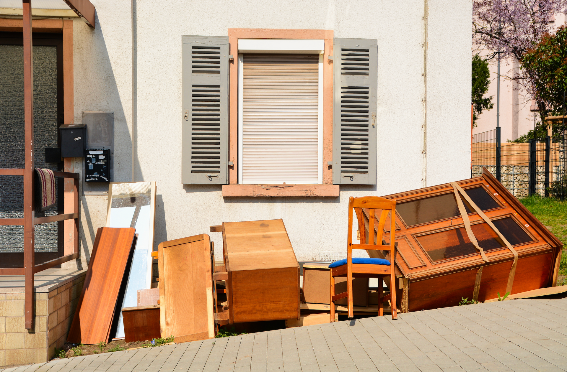 Pile of wooden furniture sits outside a white building with an open window and closed shutters.