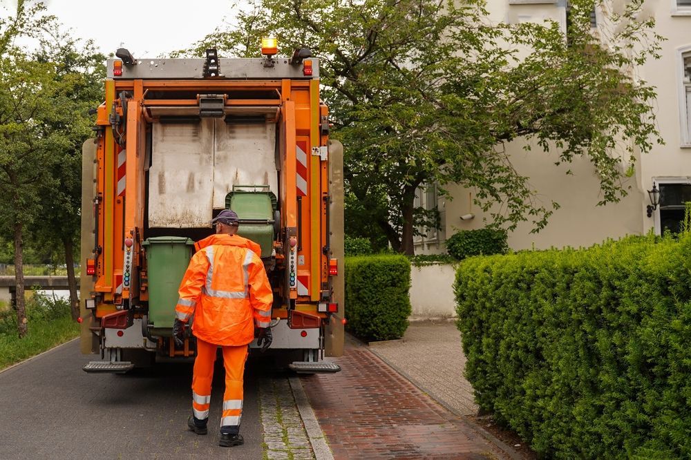 Garbage truck worker in orange suit loading green trash bins.