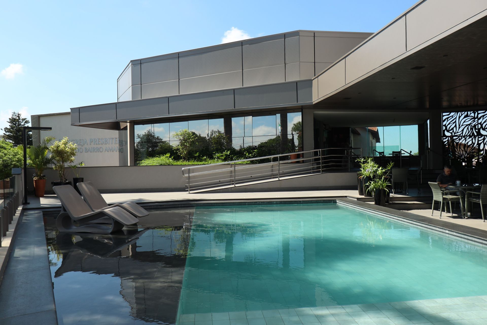 Piscina moderna na cobertura com espreguiçadeiras, guarda-corpo de vidro e um edifício contemporâneo sob um céu azul.