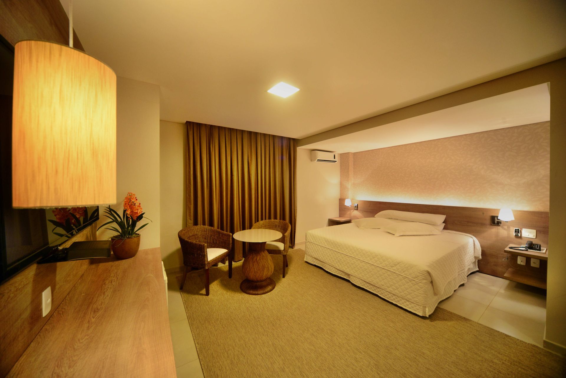 Quarto de hotel moderno com cama, duas cadeiras, iluminação aconchegante e cortinas.