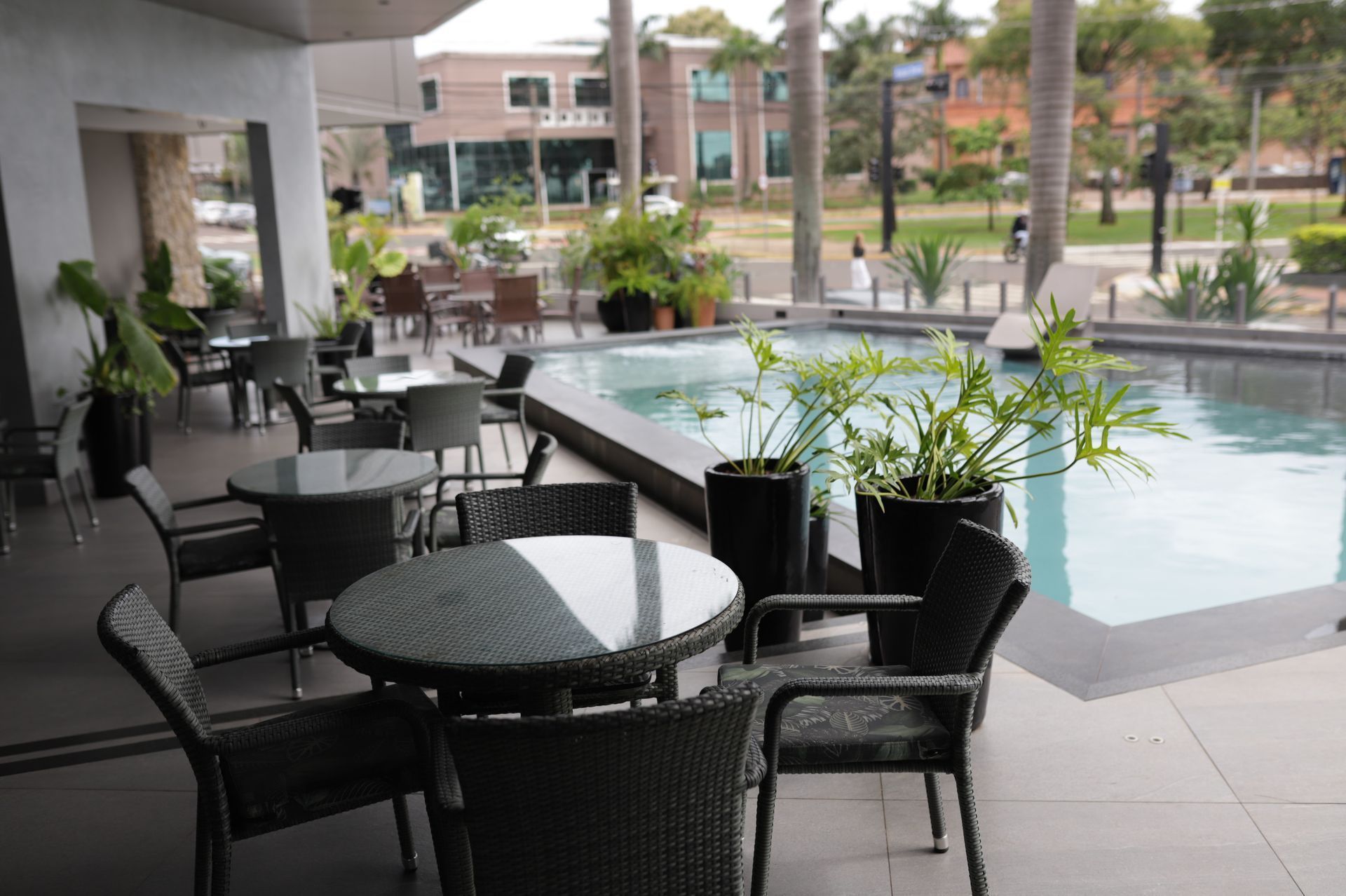 Pátio externo junto à piscina com cadeiras de vime pretas, mesas e vasos de plantas ao lado de uma piscina no pátio.