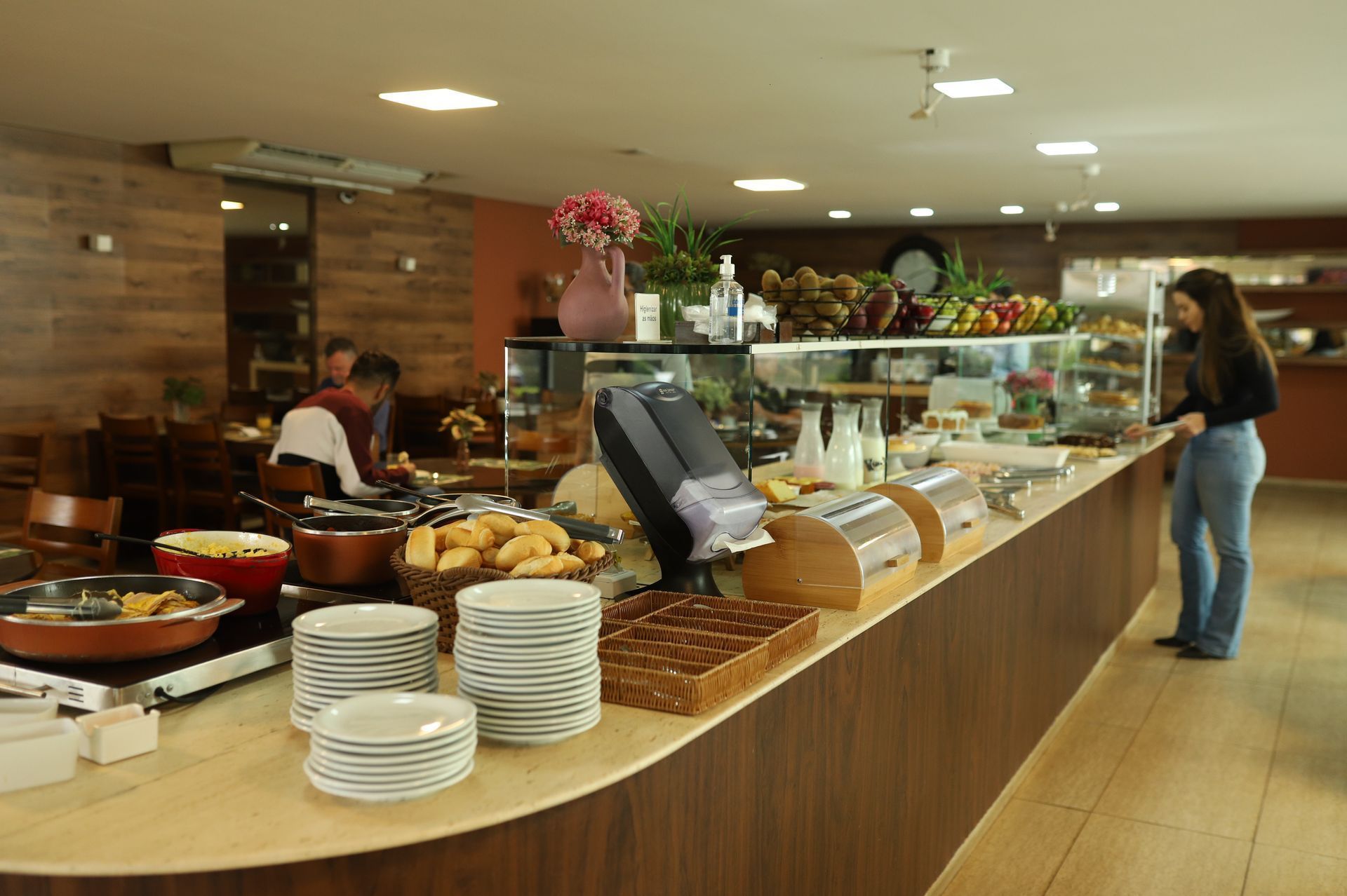 Balcão de buffet com bandejas de comida, pratos e uma mulher em pé em uma área de refeições estilo cafeteria.