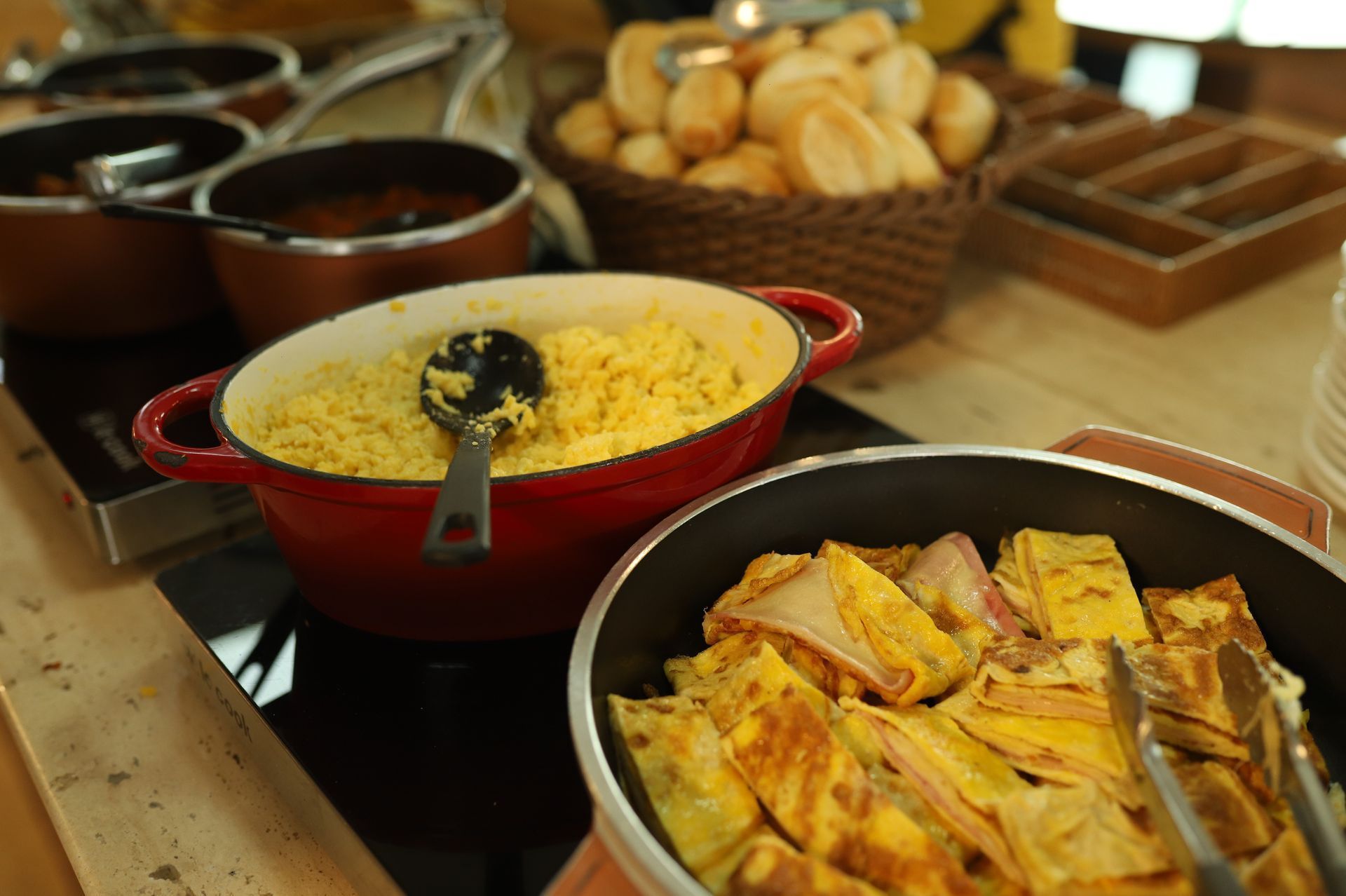 Mesa de buffet com panelas vermelhas e pretas contendo arroz, pratos assados ​​e cestas de pão.