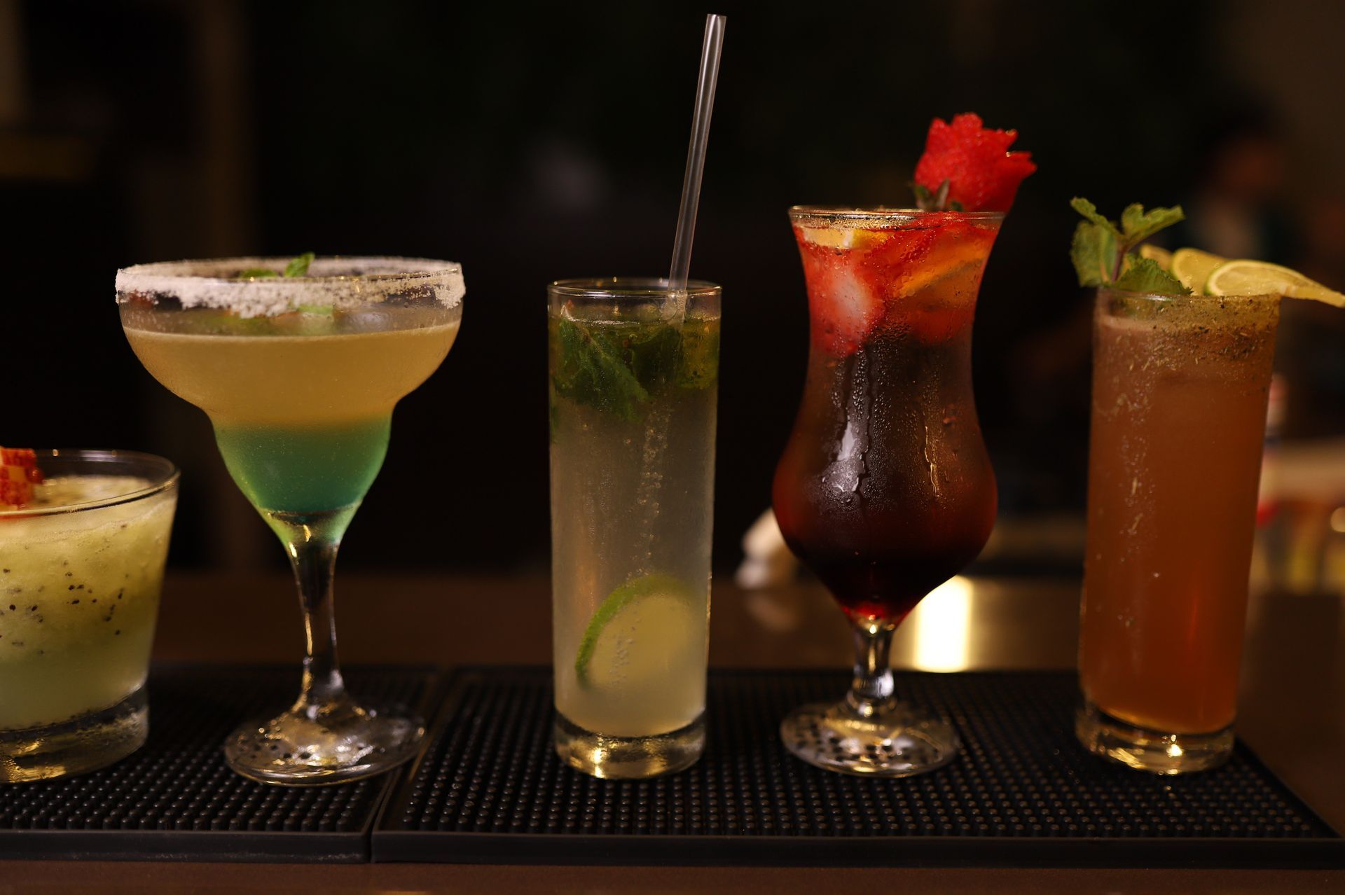 Cinco coquetéis coloridos em copos variados, enfileirados em um bar, incluindo guarnições e canudos.