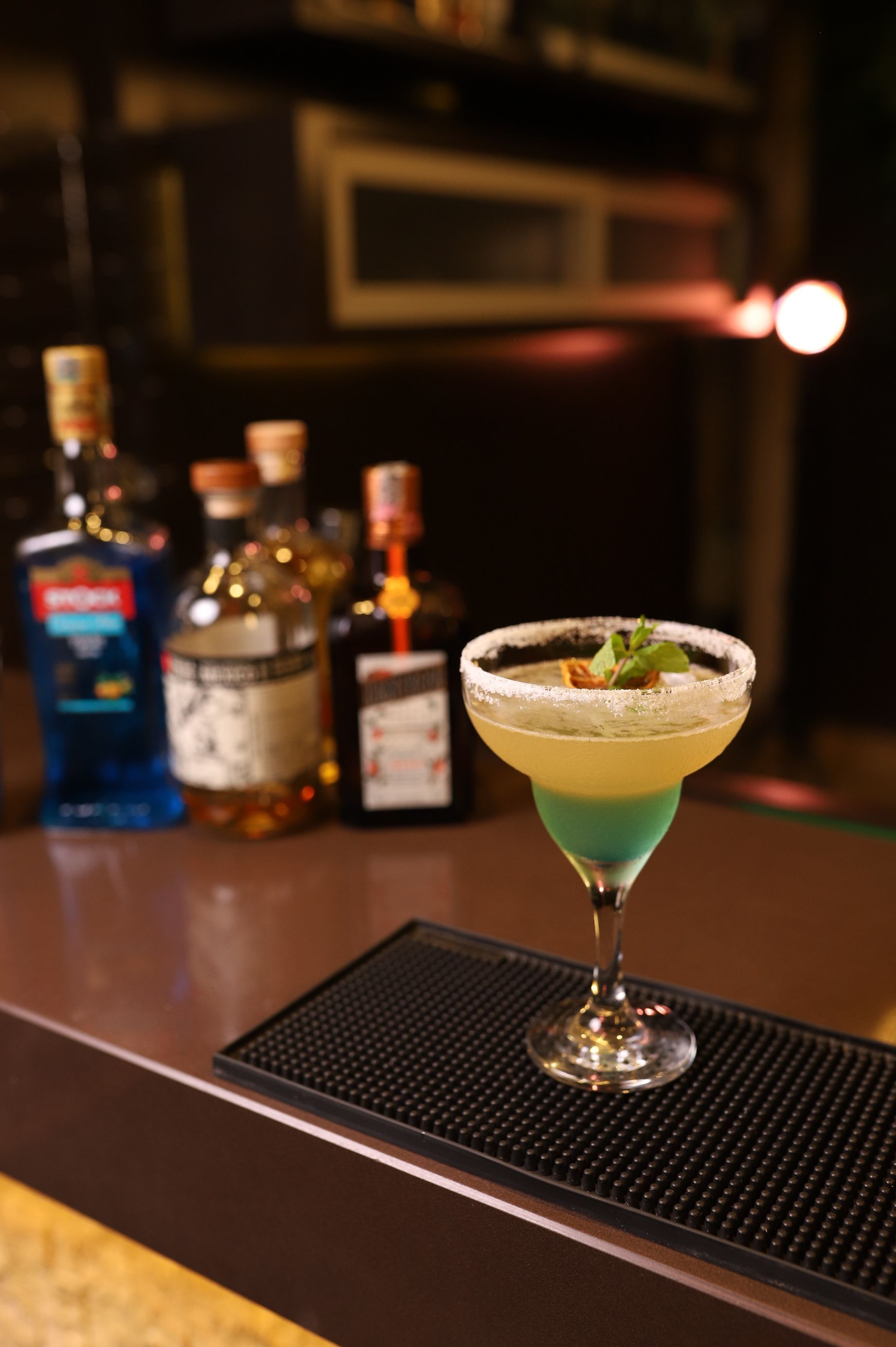 Coquetel verde em uma taça de martini sobre um bar, com garrafas de bebidas alcoólicas desfocadas ao fundo.
