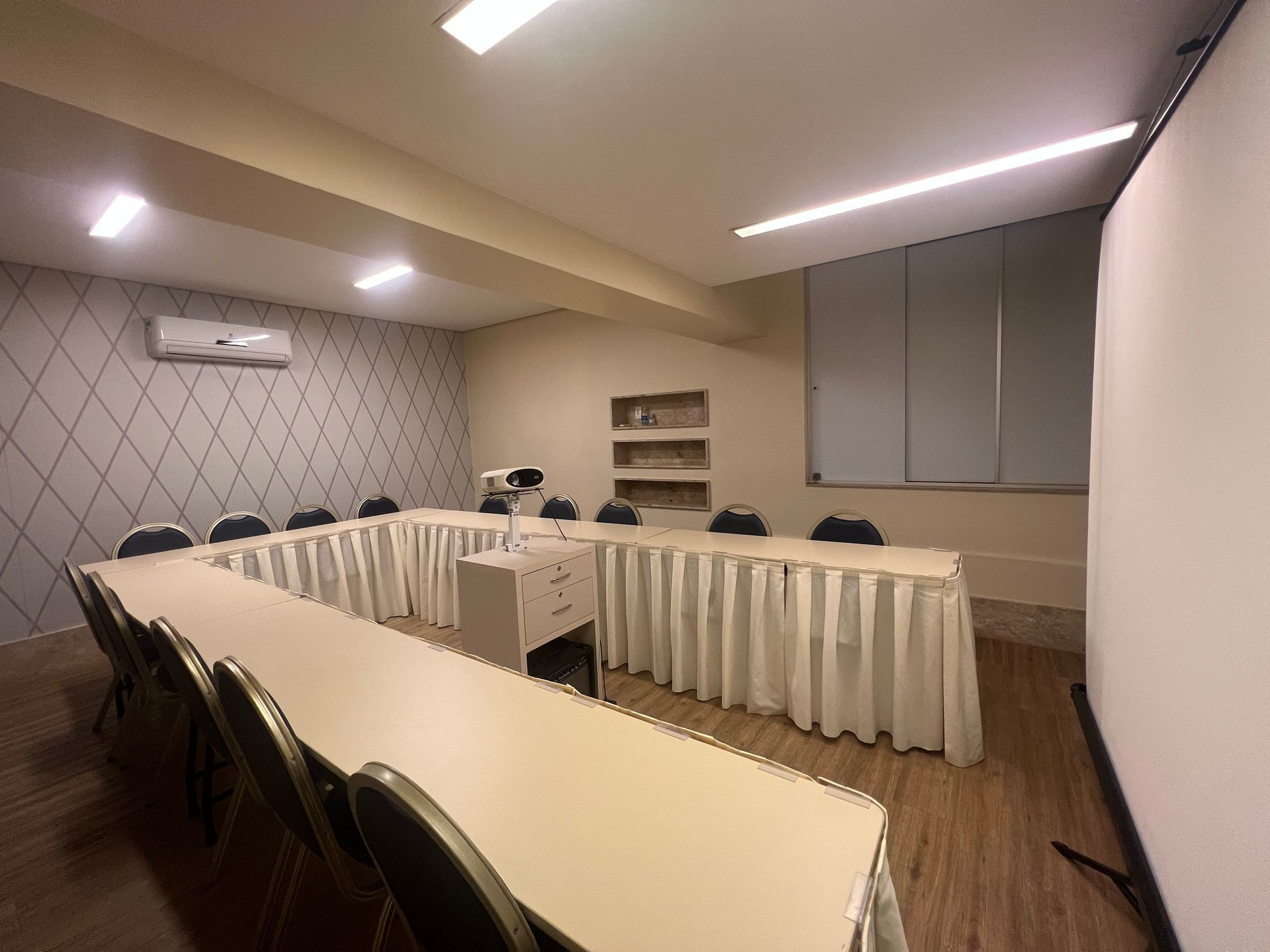 Mesa de conferência em formato de U em uma pequena sala de reuniões com toalhas de mesa brancas, cadeiras escuras e armários embutidos na parede.